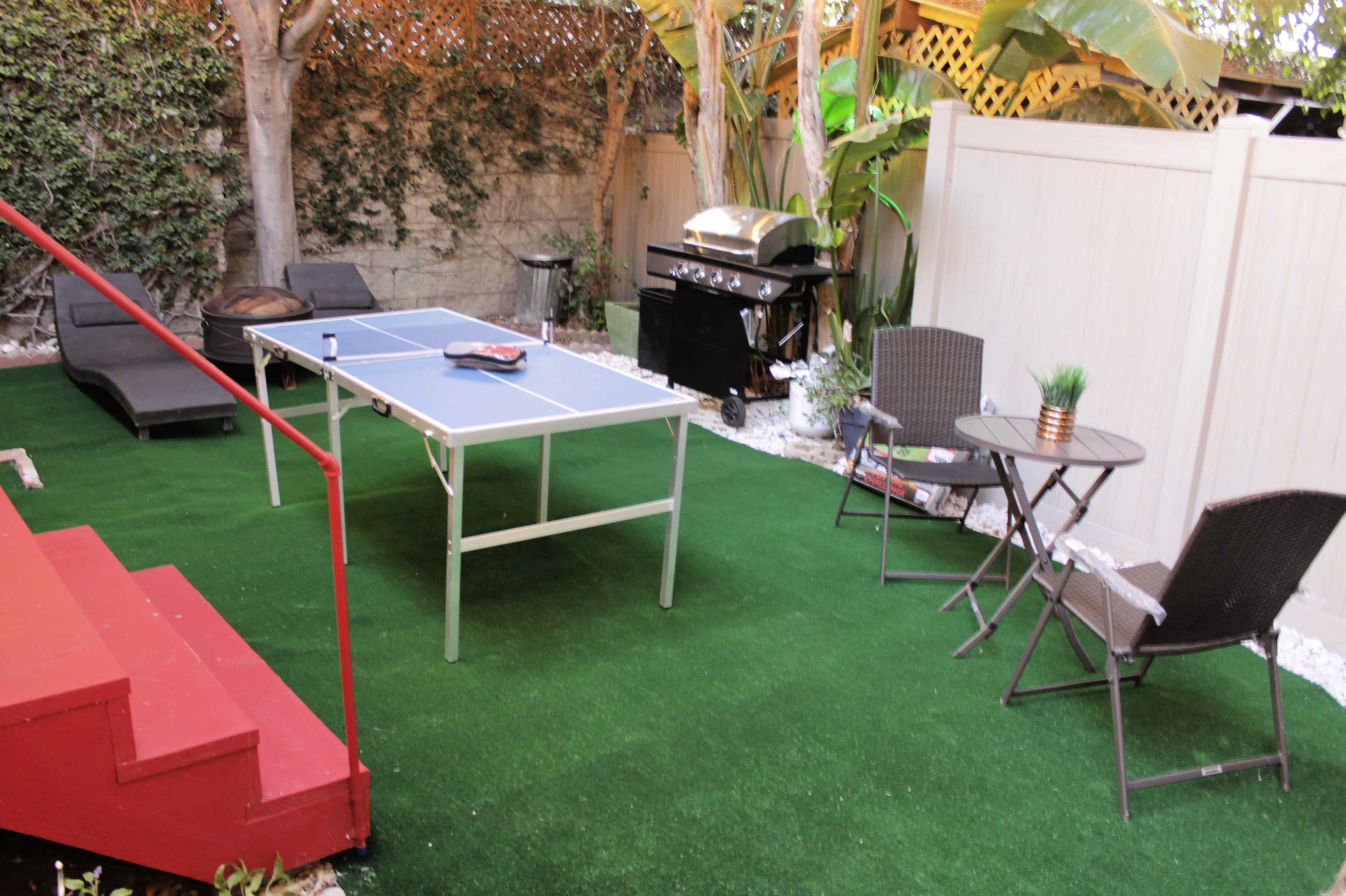 A backyard area featuring a ping pong table on artificial grass, a barbecue grill, a small dining table with chairs, and a set of stairs.