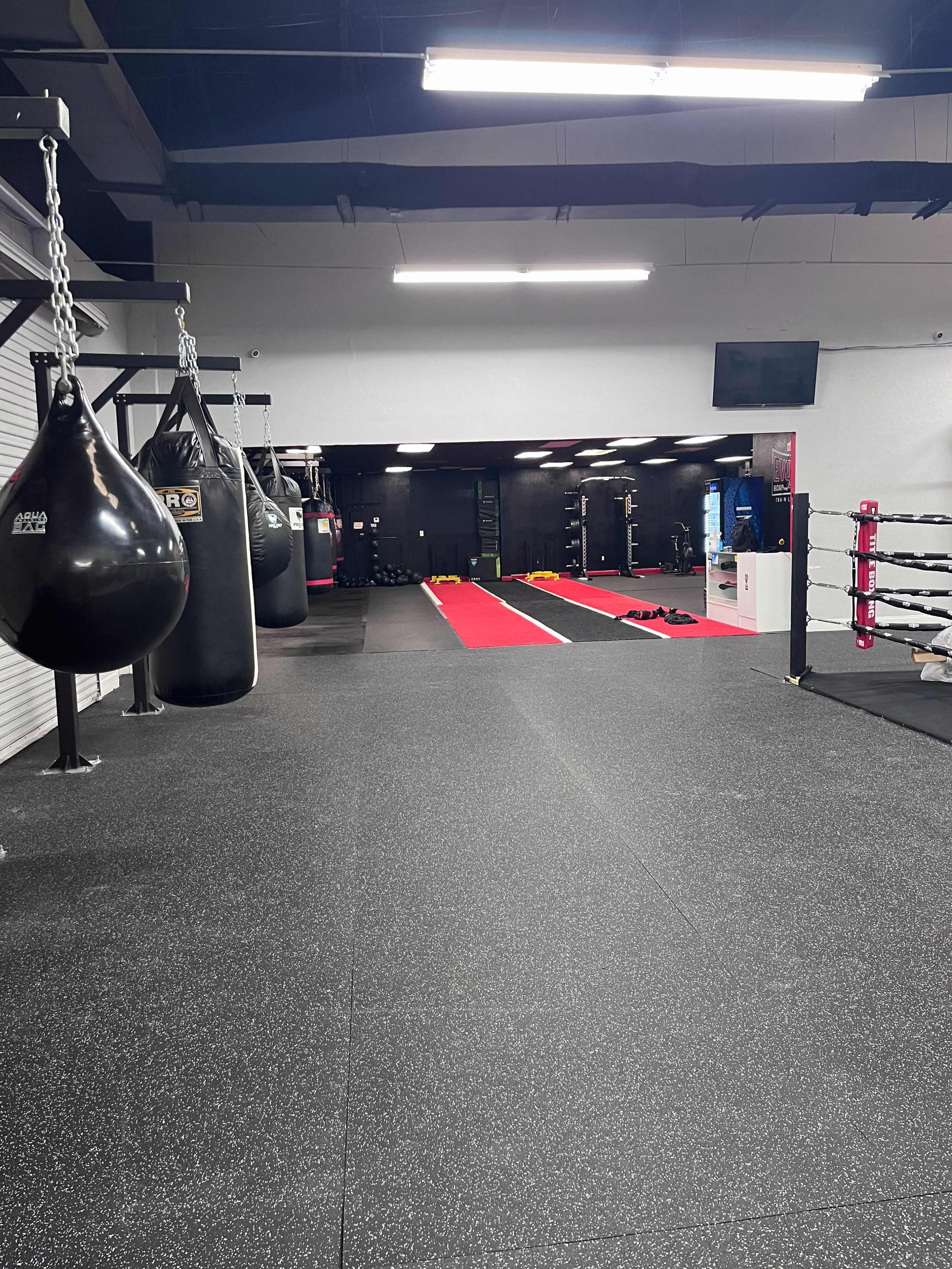 A spacious gym interior with hanging punching bags on one side and a training area featuring red mats and various fitness equipment on the other.