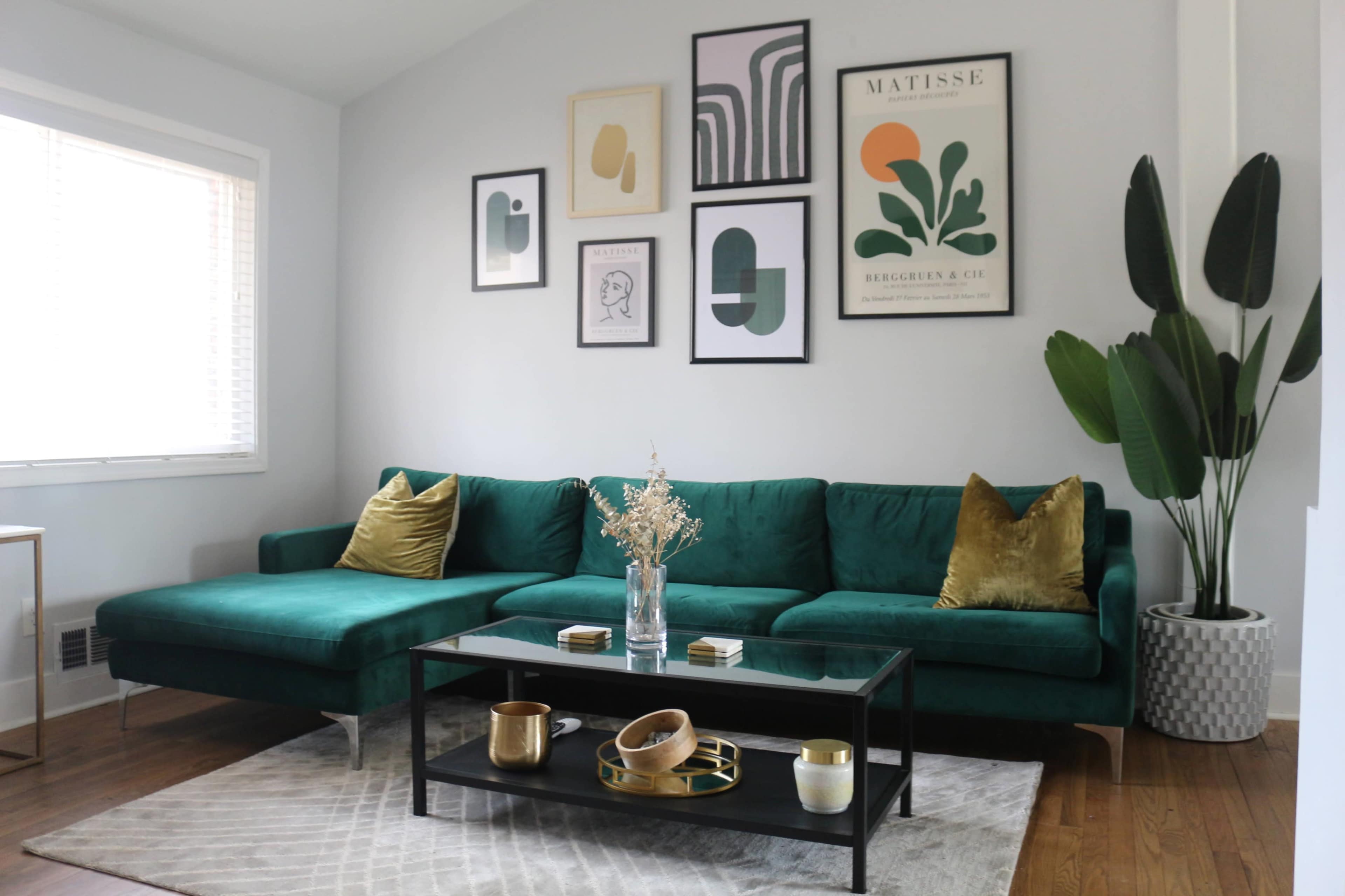 The image shows a modern living room featuring a green velvet sectional sofa, a black coffee table with decorative items, and framed art on the walls.
