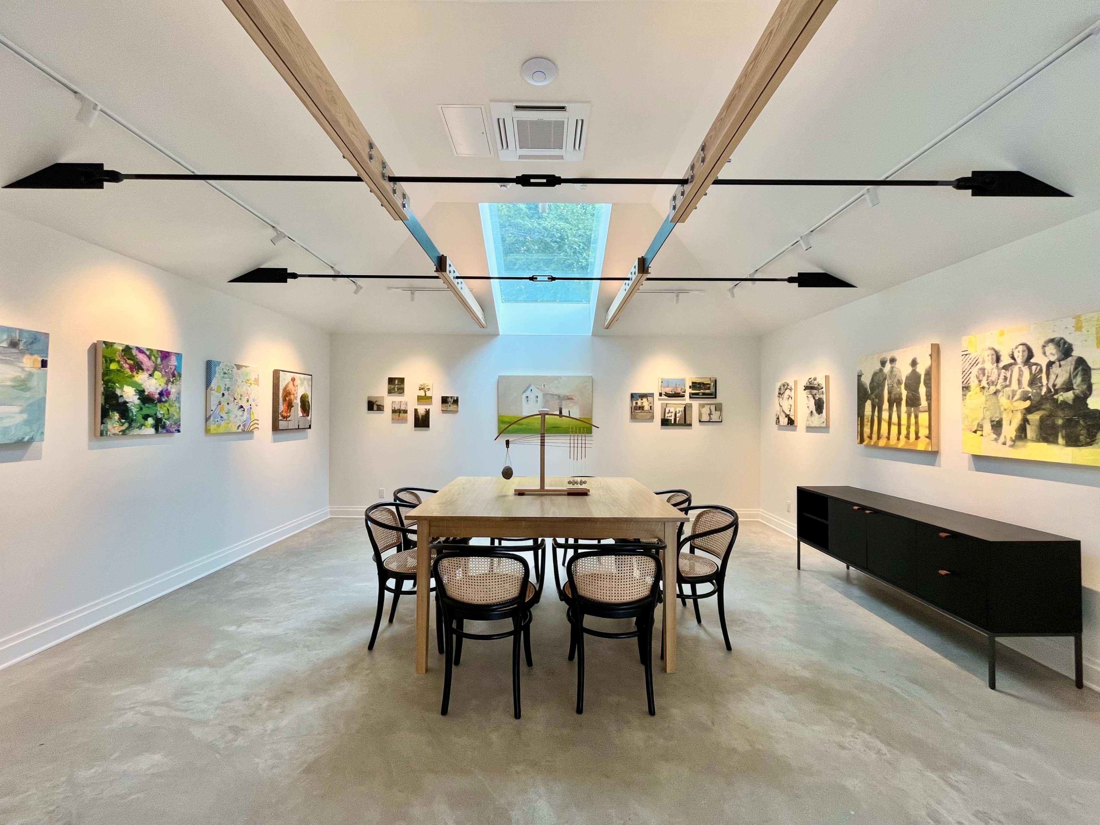 A modern art gallery features a long wooden table surrounded by six chairs, with various artworks displayed on white walls and a skylight providing natural light.