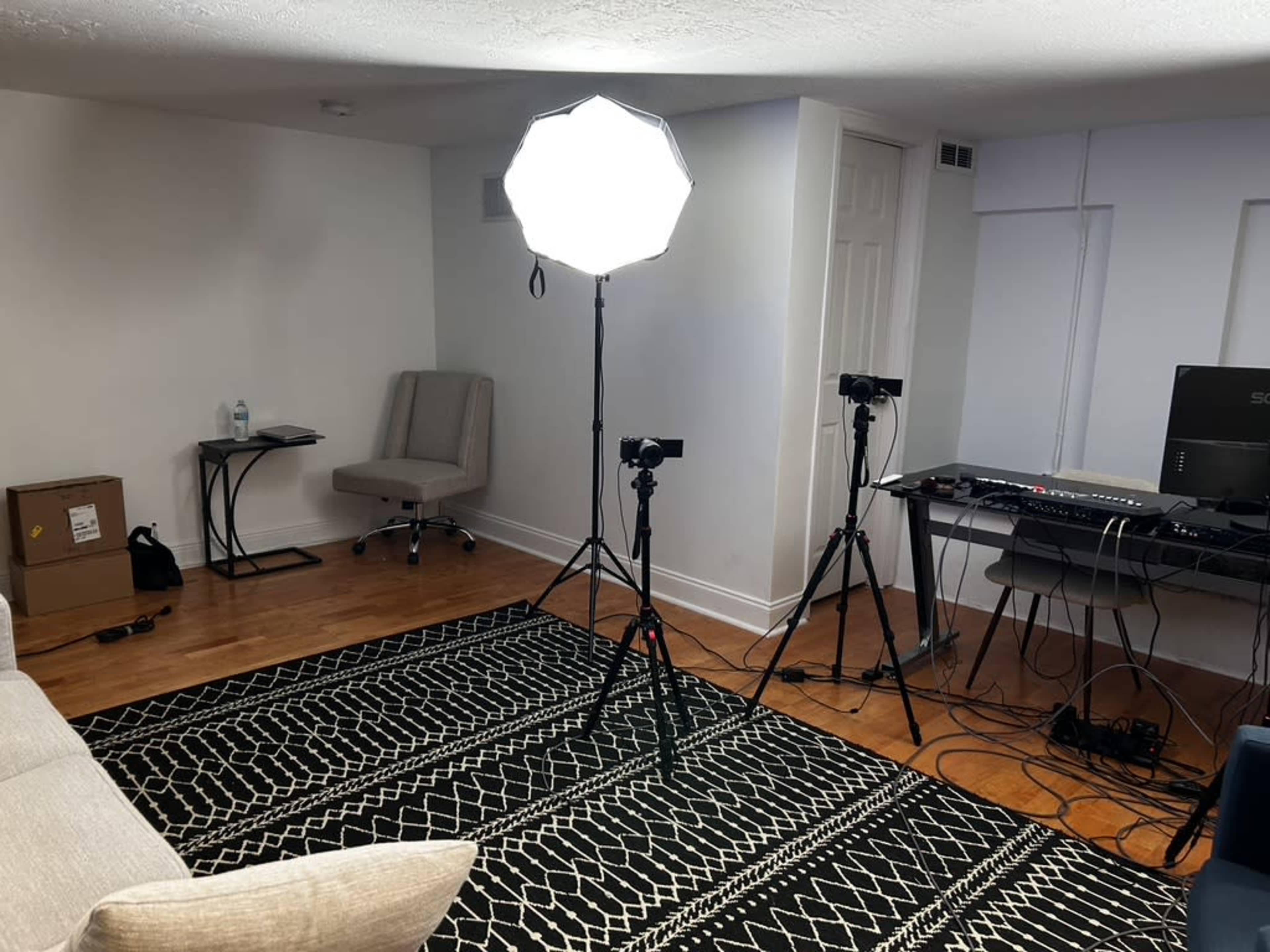 A well-lit room features a sofa, a small table, two cameras on tripods, a keyboard setup, and a patterned rug.