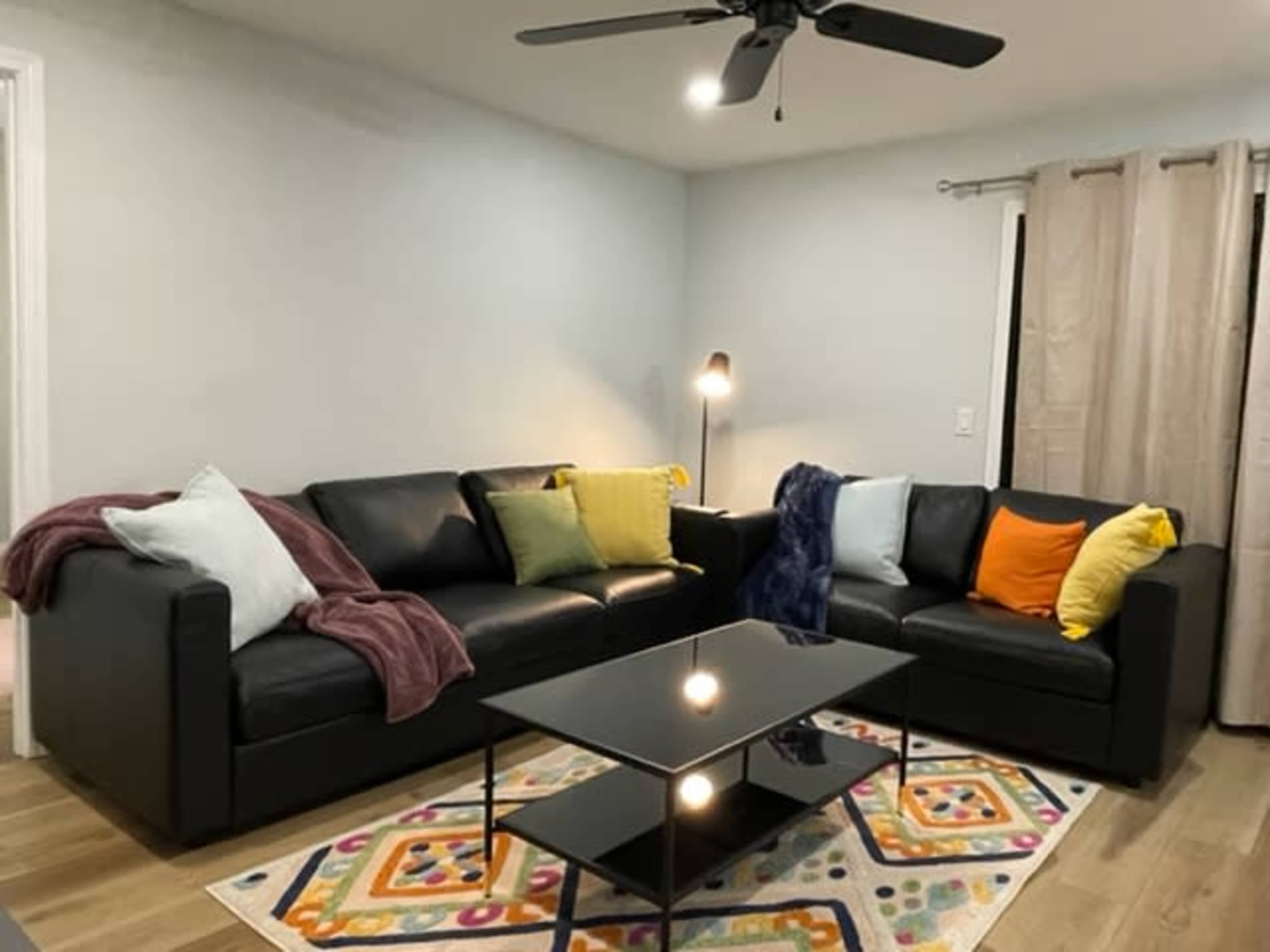 The room features a black leather sectional sofa adorned with colorful pillows and a coffee table placed on a patterned area rug.