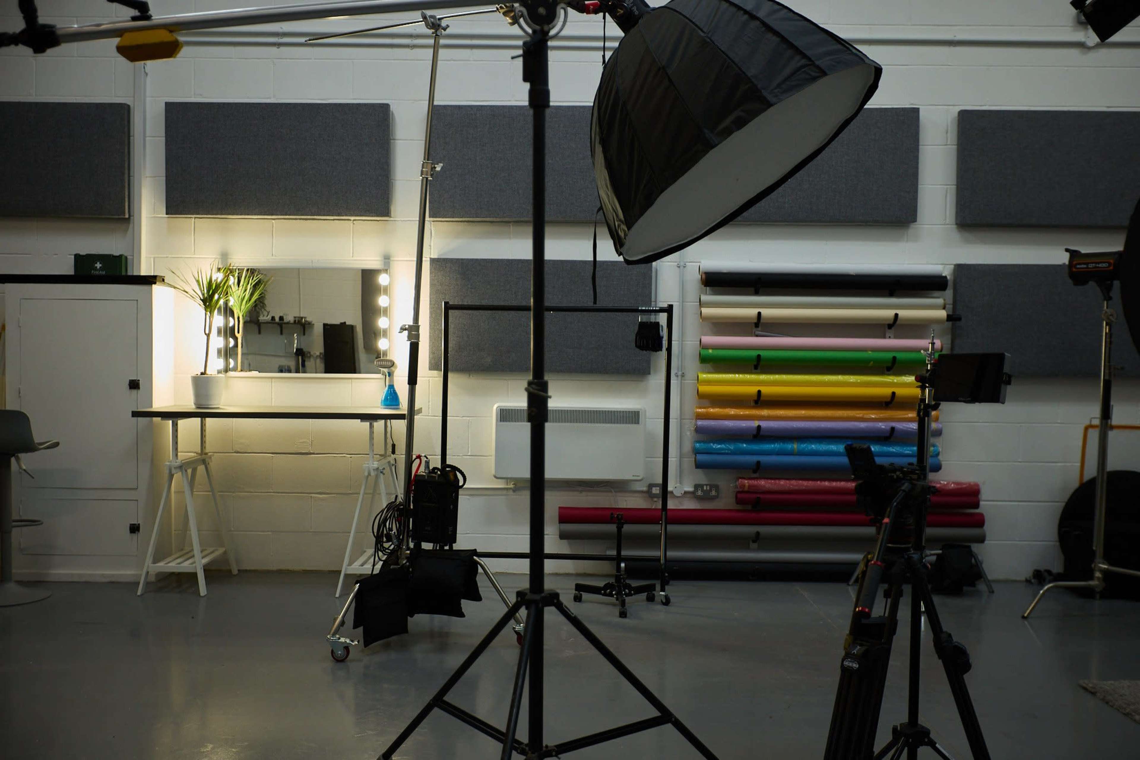 A photography studio featuring a light setup, a mirror with bulbs, and a backdrop rack with colorful roll paper.
