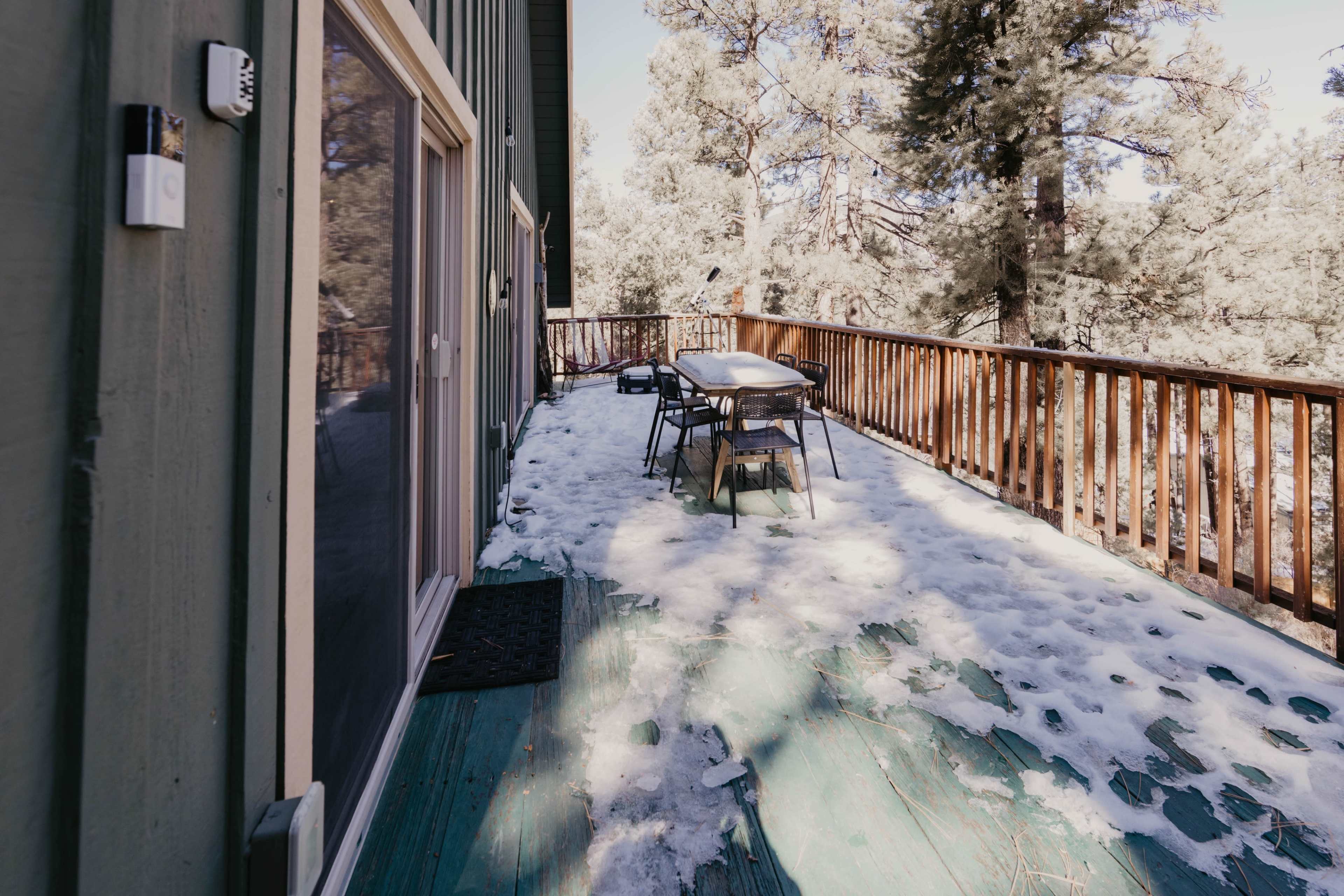 A deck covered with snow and surrounded by pine trees is shown, with a table and chairs positioned on the surface.