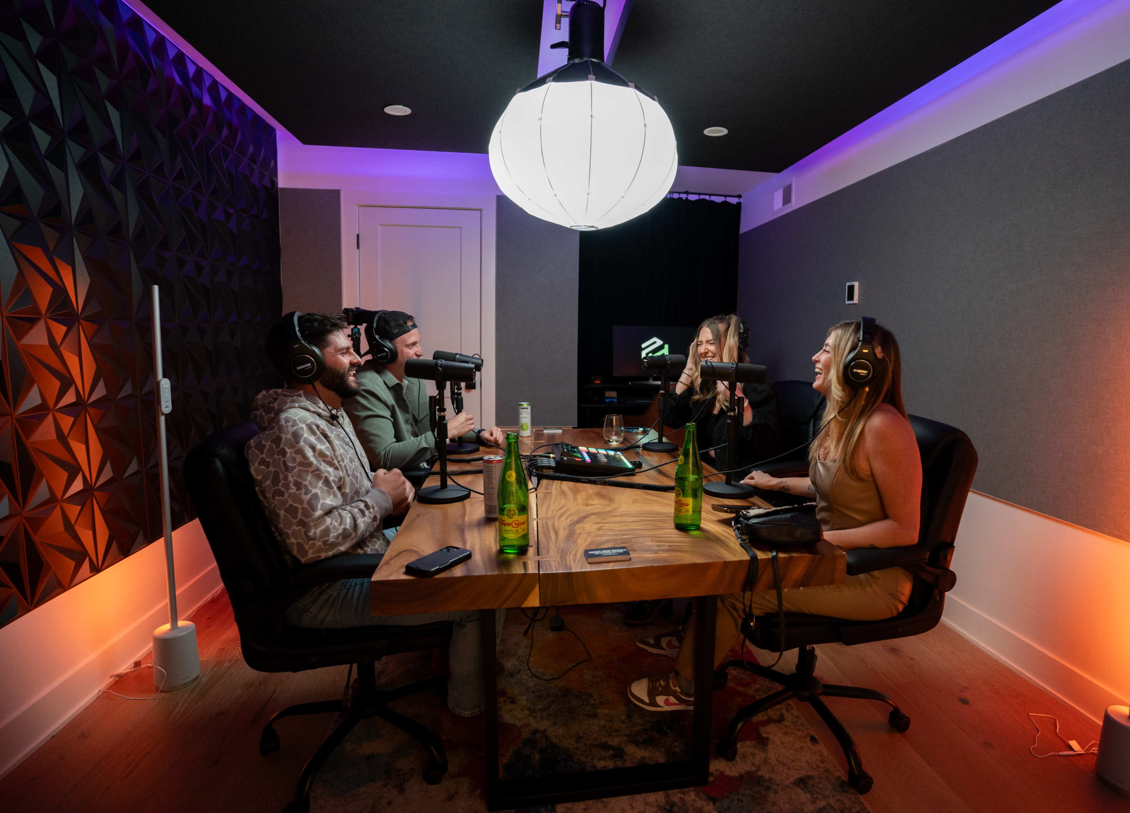 Four people sit around a table in a podcast studio, equipped with microphones and drinks, engaged in conversation under a large overhead light.