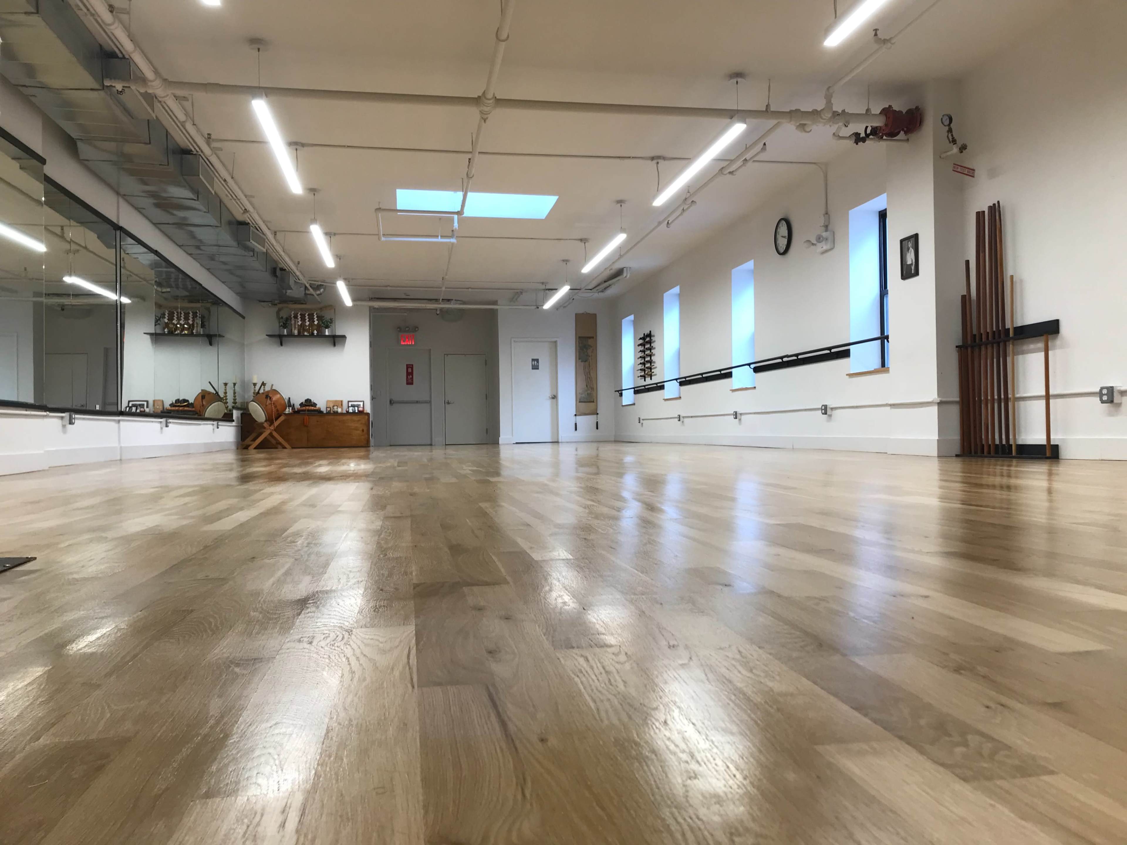 The image shows an empty dance studio with wooden flooring, mirrors along one wall, and bright overhead lighting.