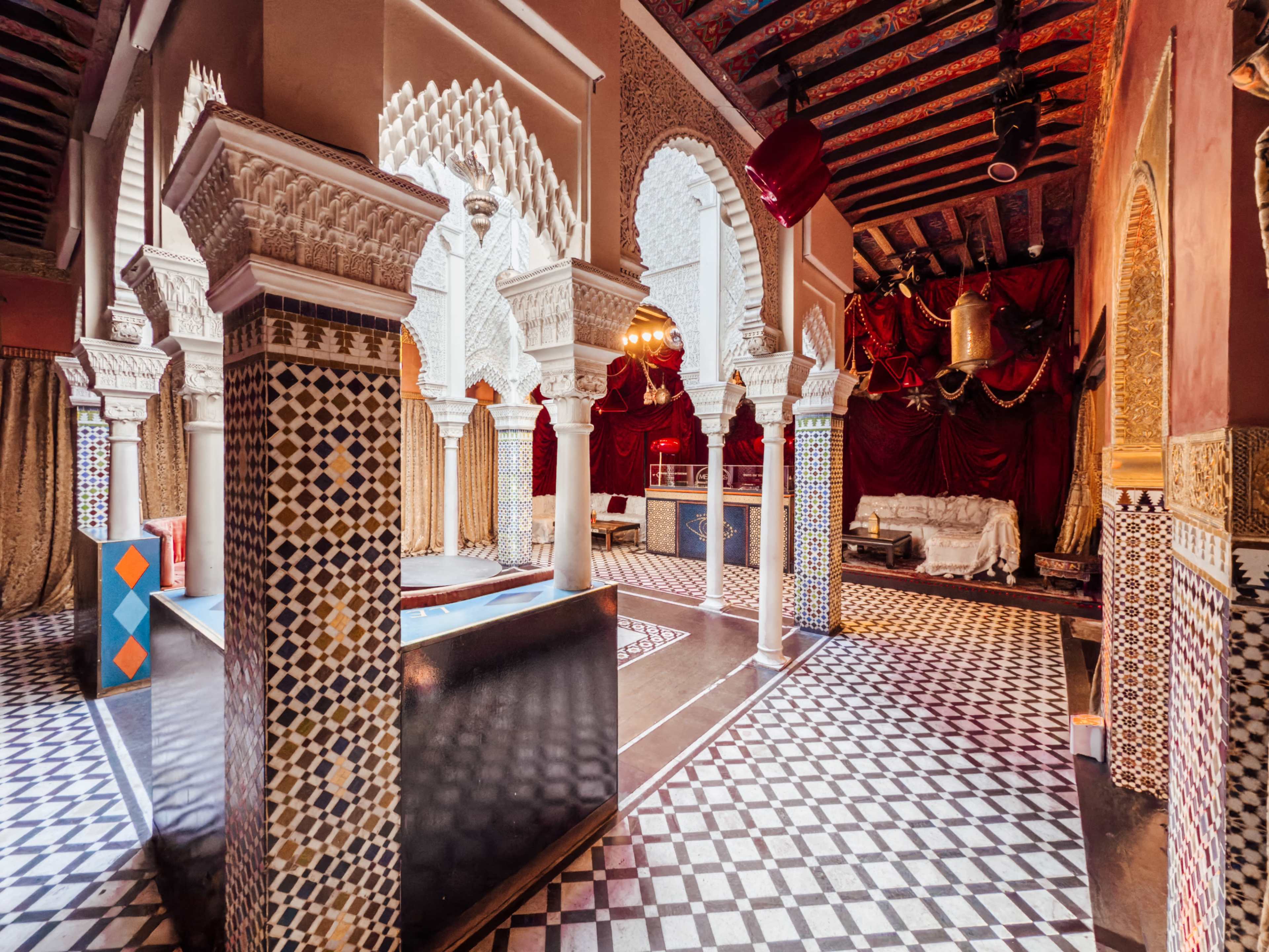 The image shows an ornate interior space featuring intricate archways, detailed tile work, and a patterned floor, characteristic of Moroccan architectural design.