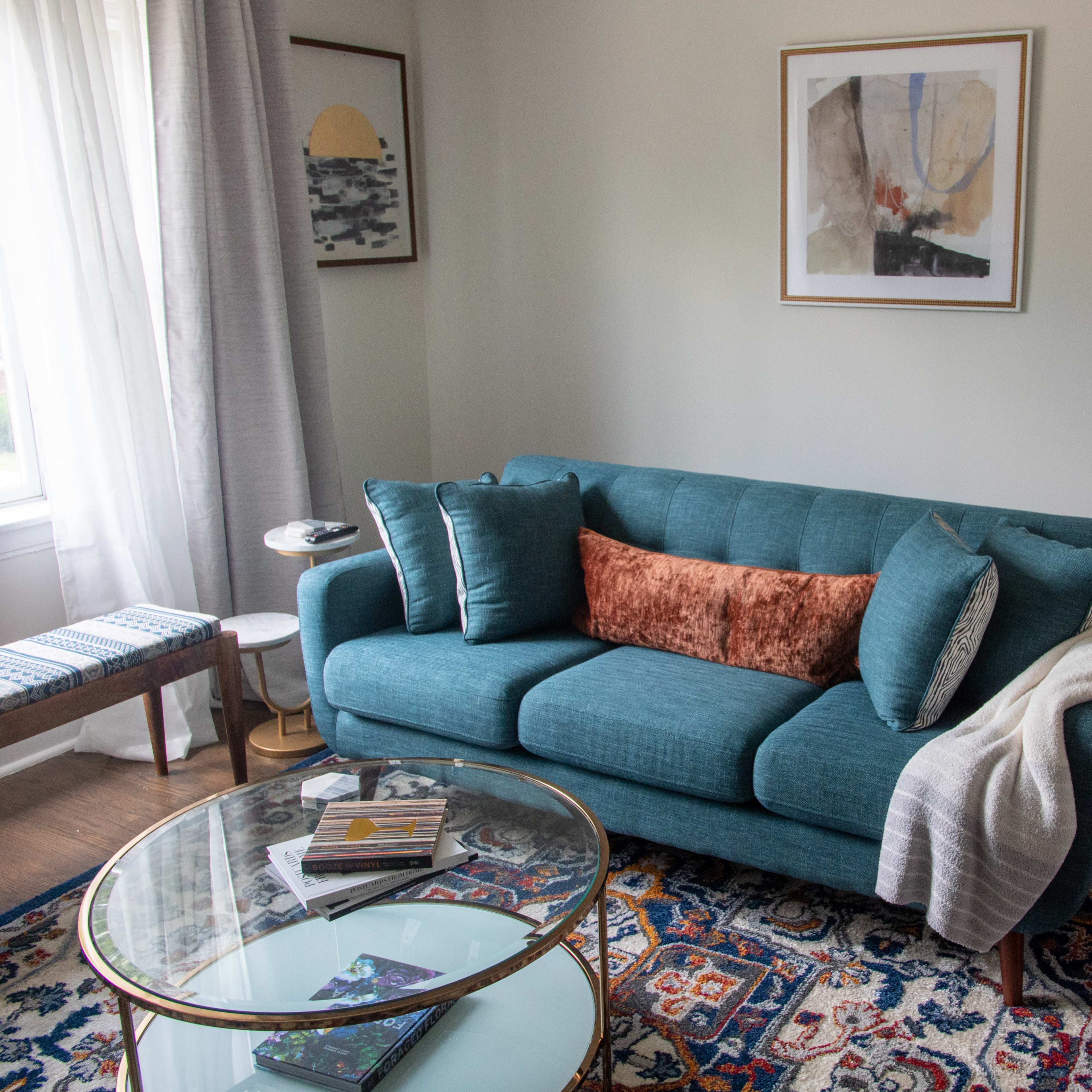 A turquoise sofa with cushions sits in a well-lit living room, accompanied by a glass coffee table and a patterned rug.