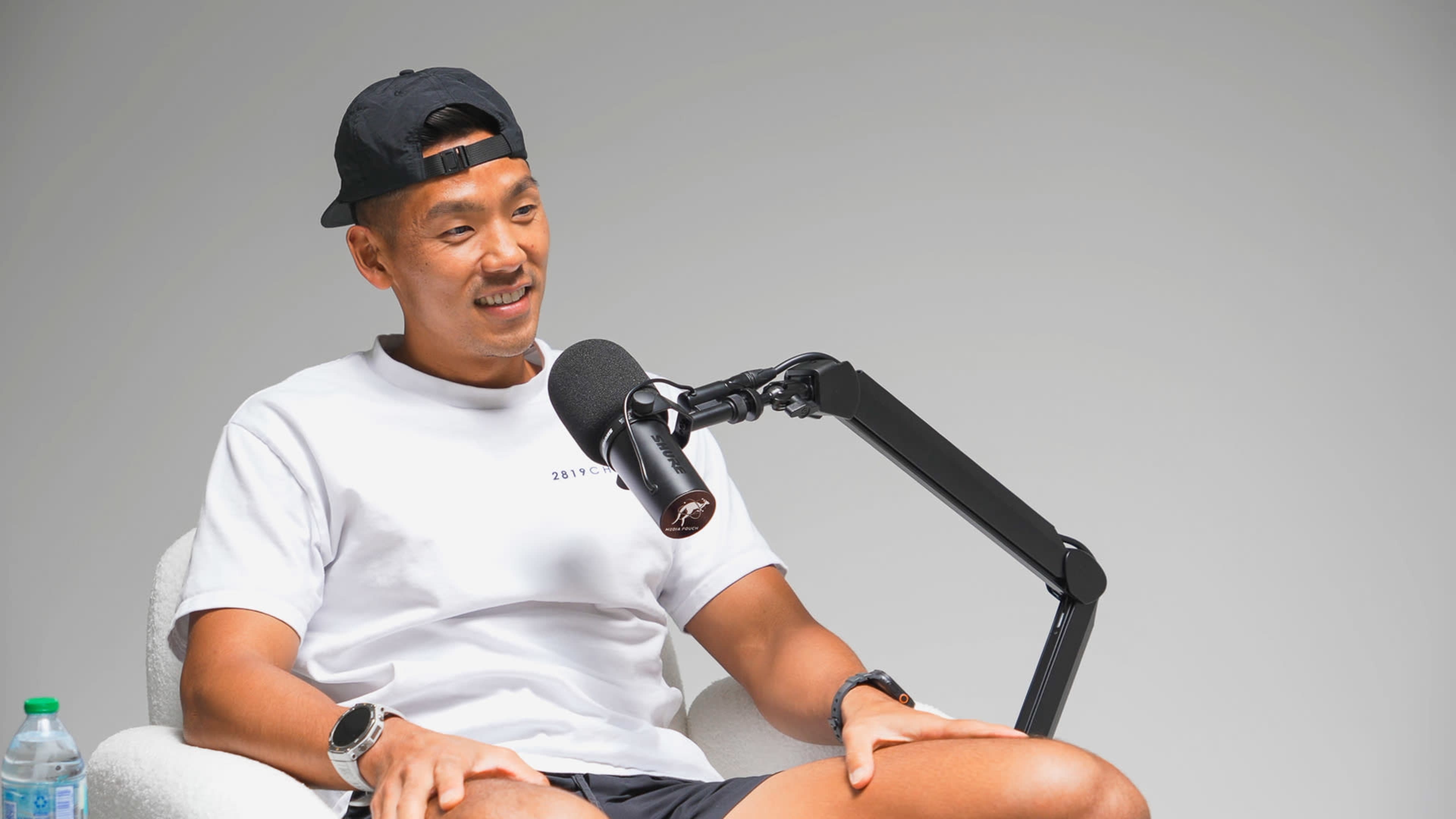 A man with short hair and a cap sits in a chair, smiling while speaking into a microphone during a podcast recording.
