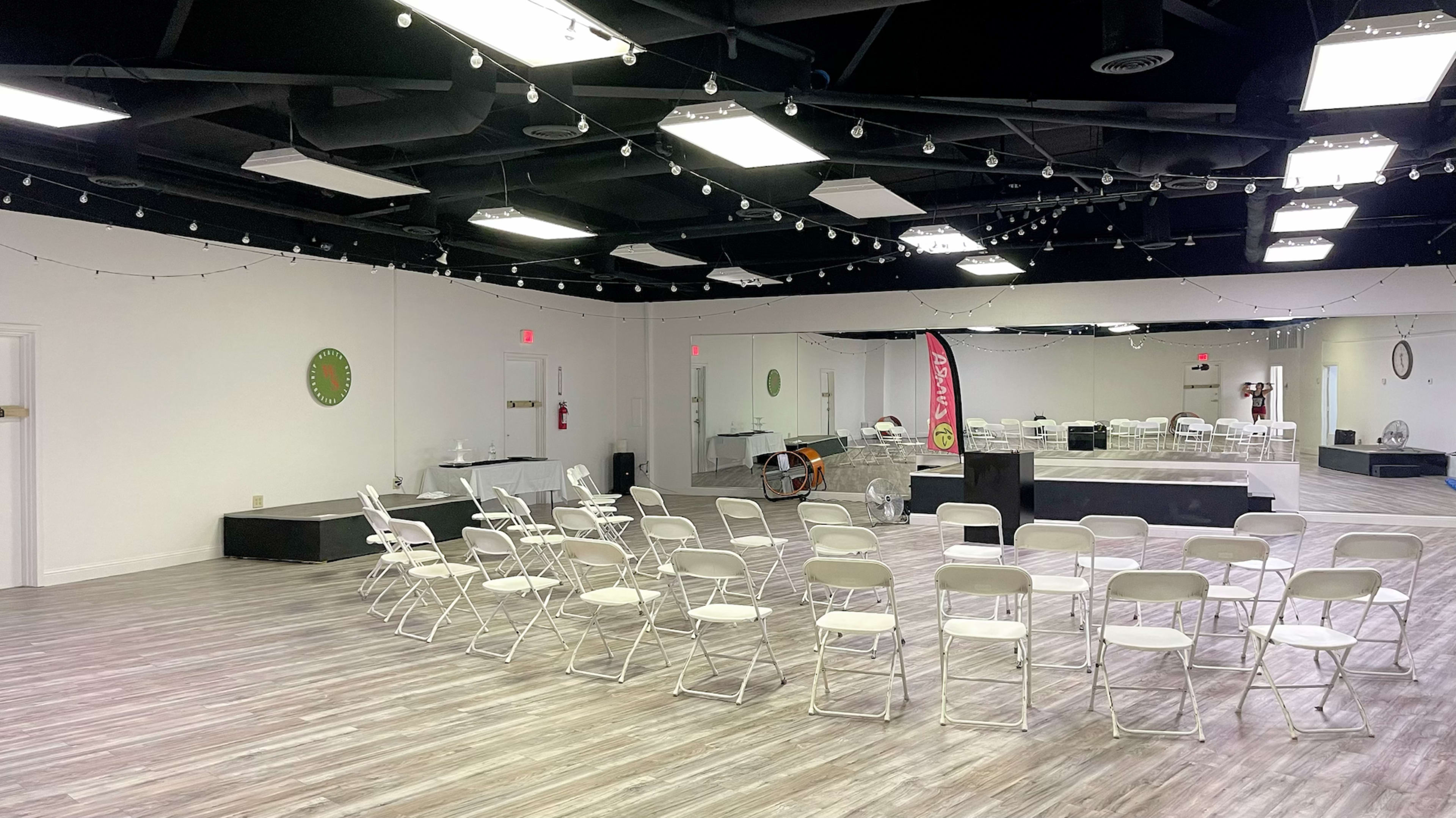 A spacious, well-lit event room with rows of white chairs facing a stage area, complemented by mirrors and decorative elements on the walls.