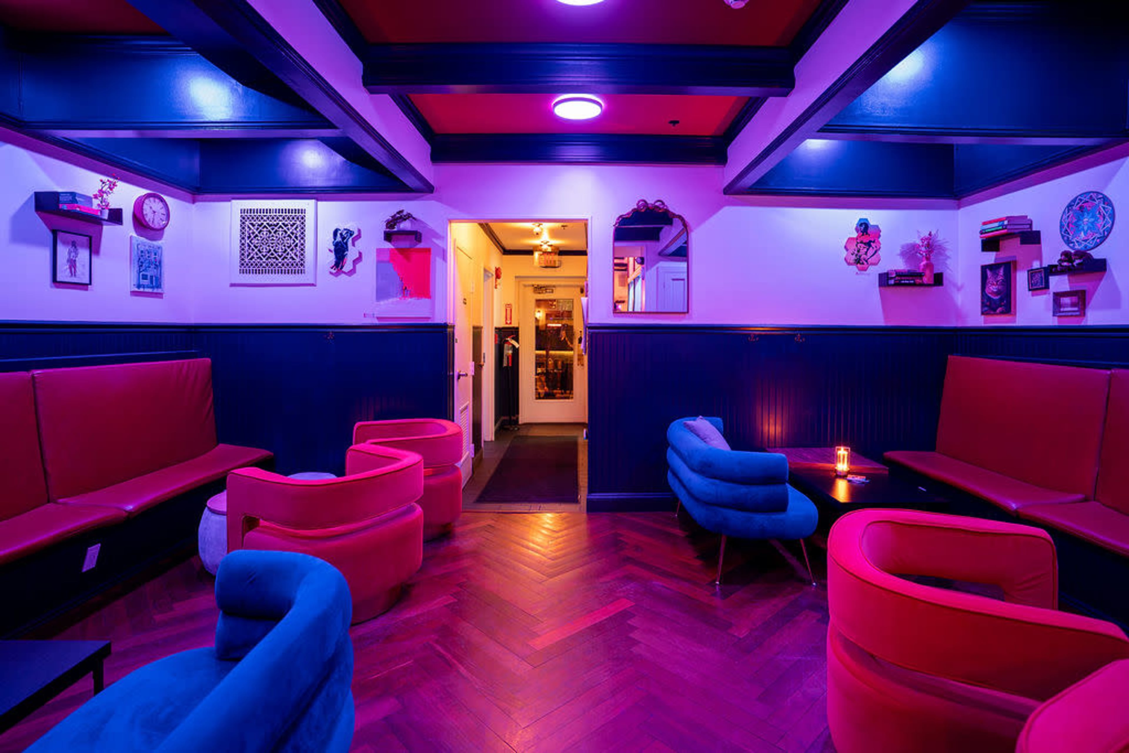 The image depicts a dimly lit lounge with colorful seating, featuring red and blue chairs arranged around small tables beneath a multi-colored ceiling.