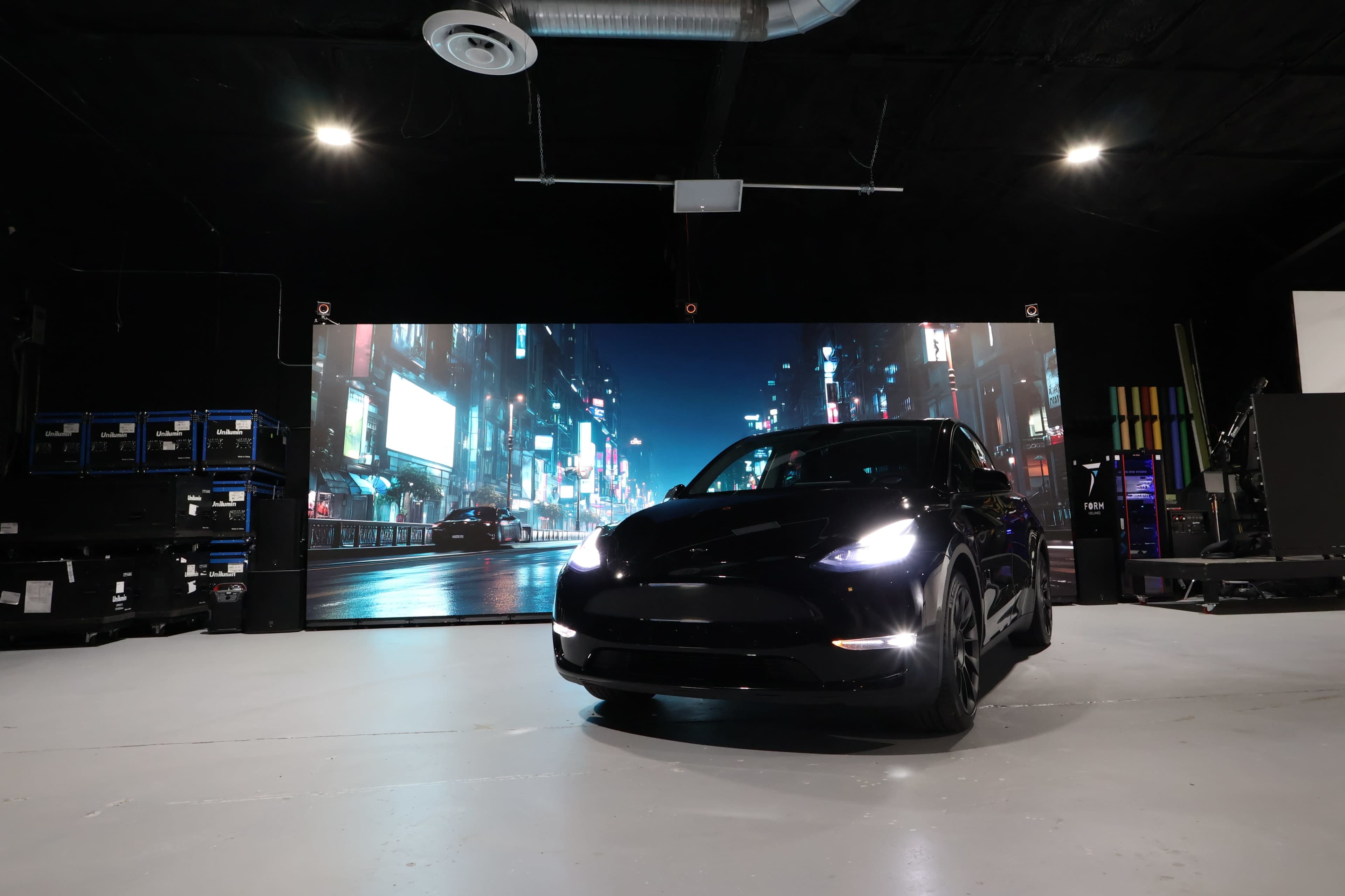 A black car is positioned in front of a large digital display showcasing a cityscape at night.