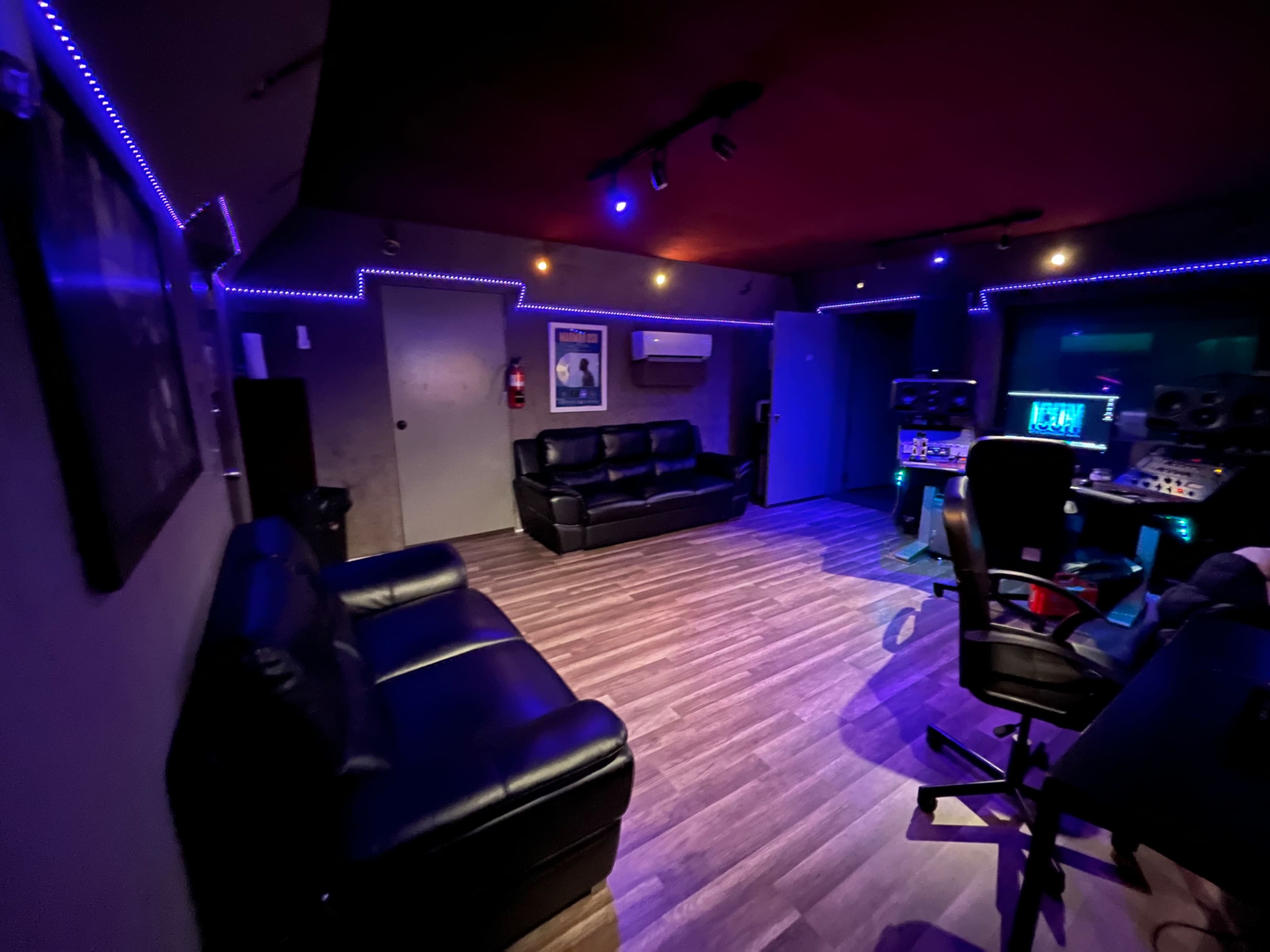 A dimly lit music studio featuring black leather sofas, a desk with studio equipment, and ambient lighting along the walls.