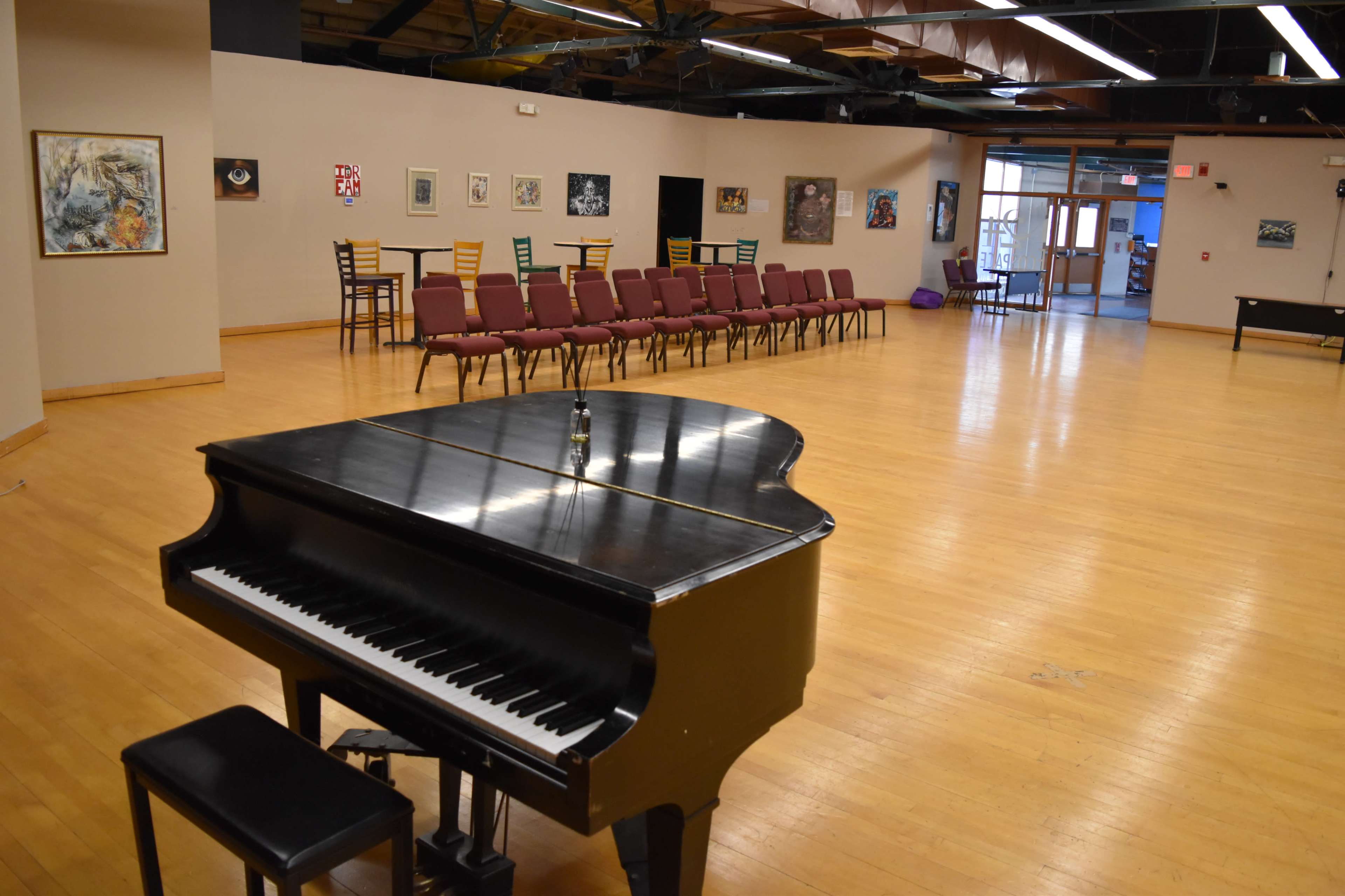 A black piano is positioned in the foreground of a spacious room with wooden flooring, which features rows of maroon chairs and various artworks displayed on the walls.