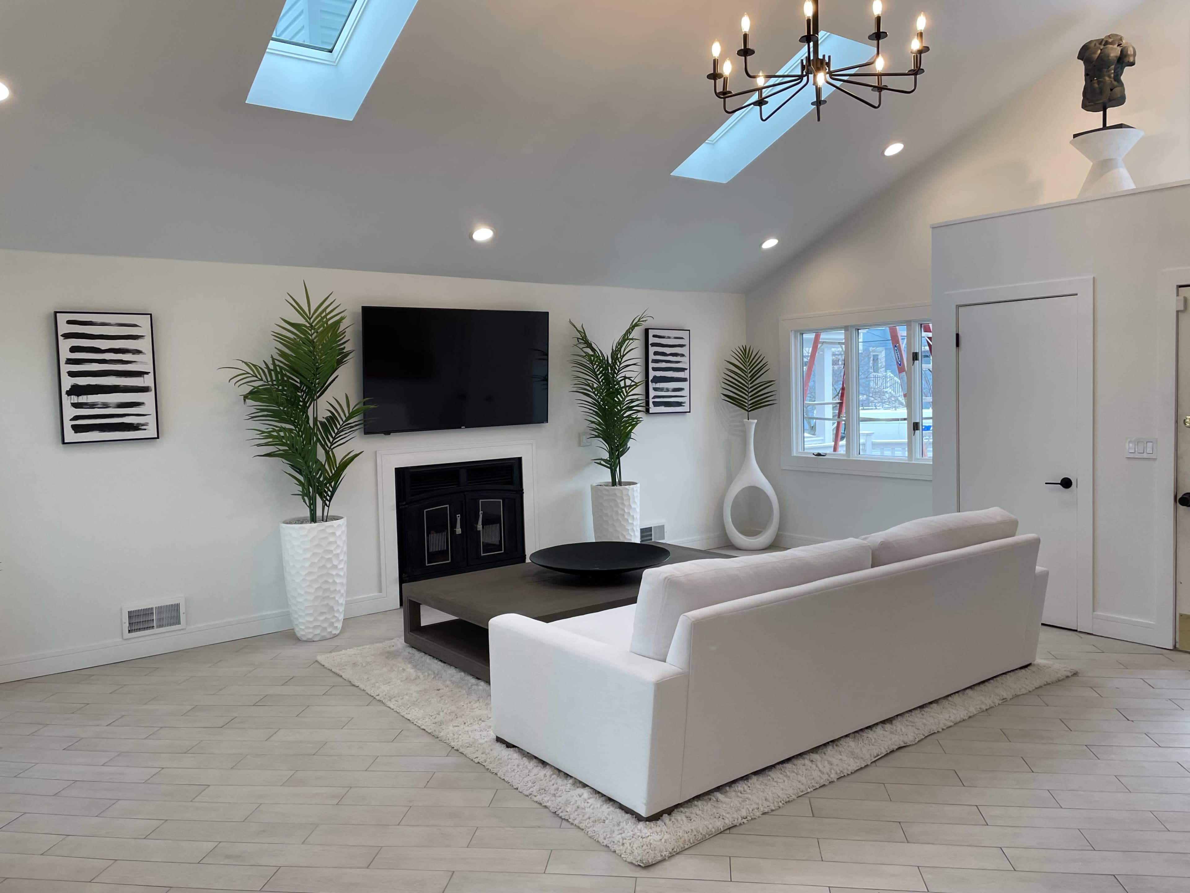 The image shows a modern living room with a white sectional couch, a dark coffee table, potted palms, and a television mounted on the wall beneath a skylight.