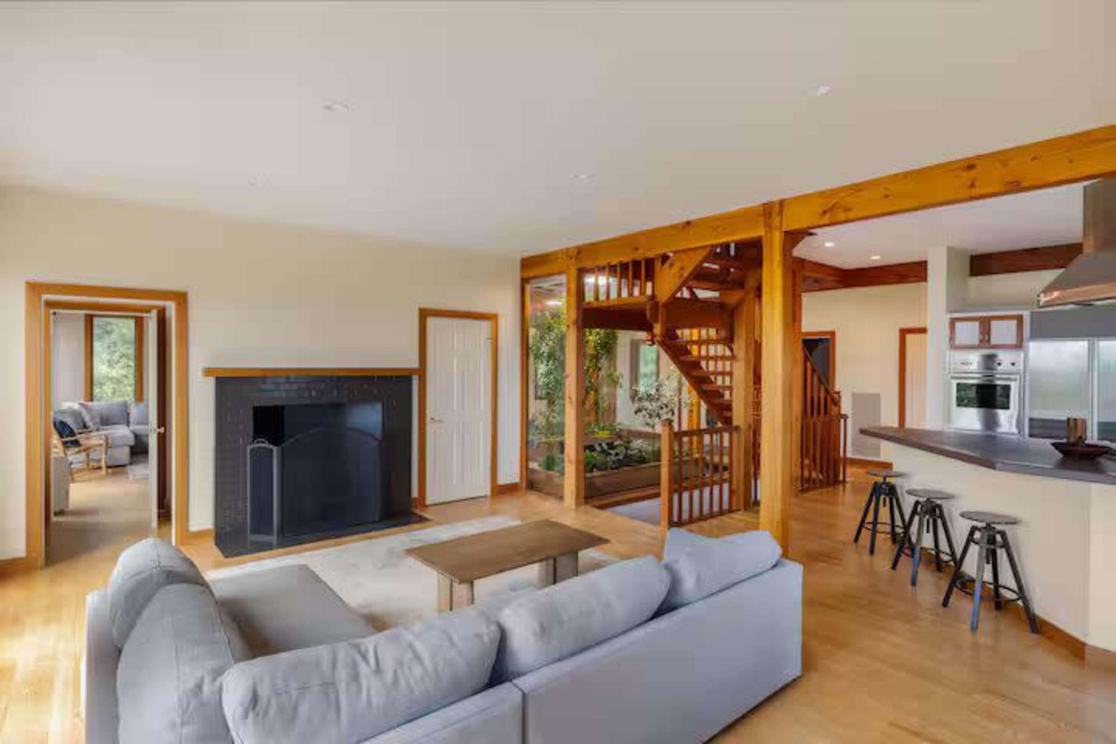 The image shows a spacious living area featuring a gray sofa, a coffee table, a fireplace, a staircase, and a kitchen with bar stools.