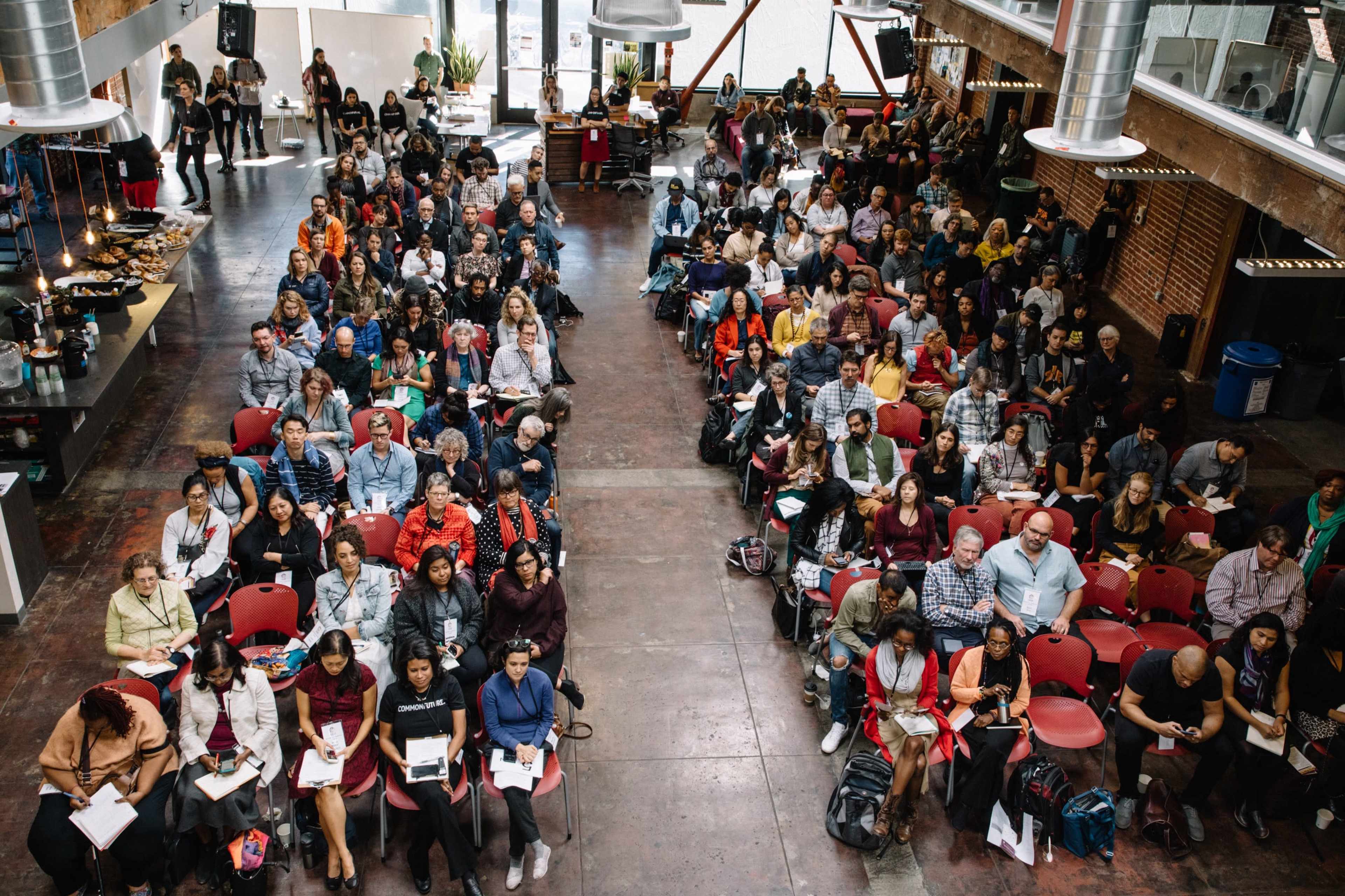 A large audience is seated in a spacious venue with a variety of individuals engaged in discussion and note-taking during an event.