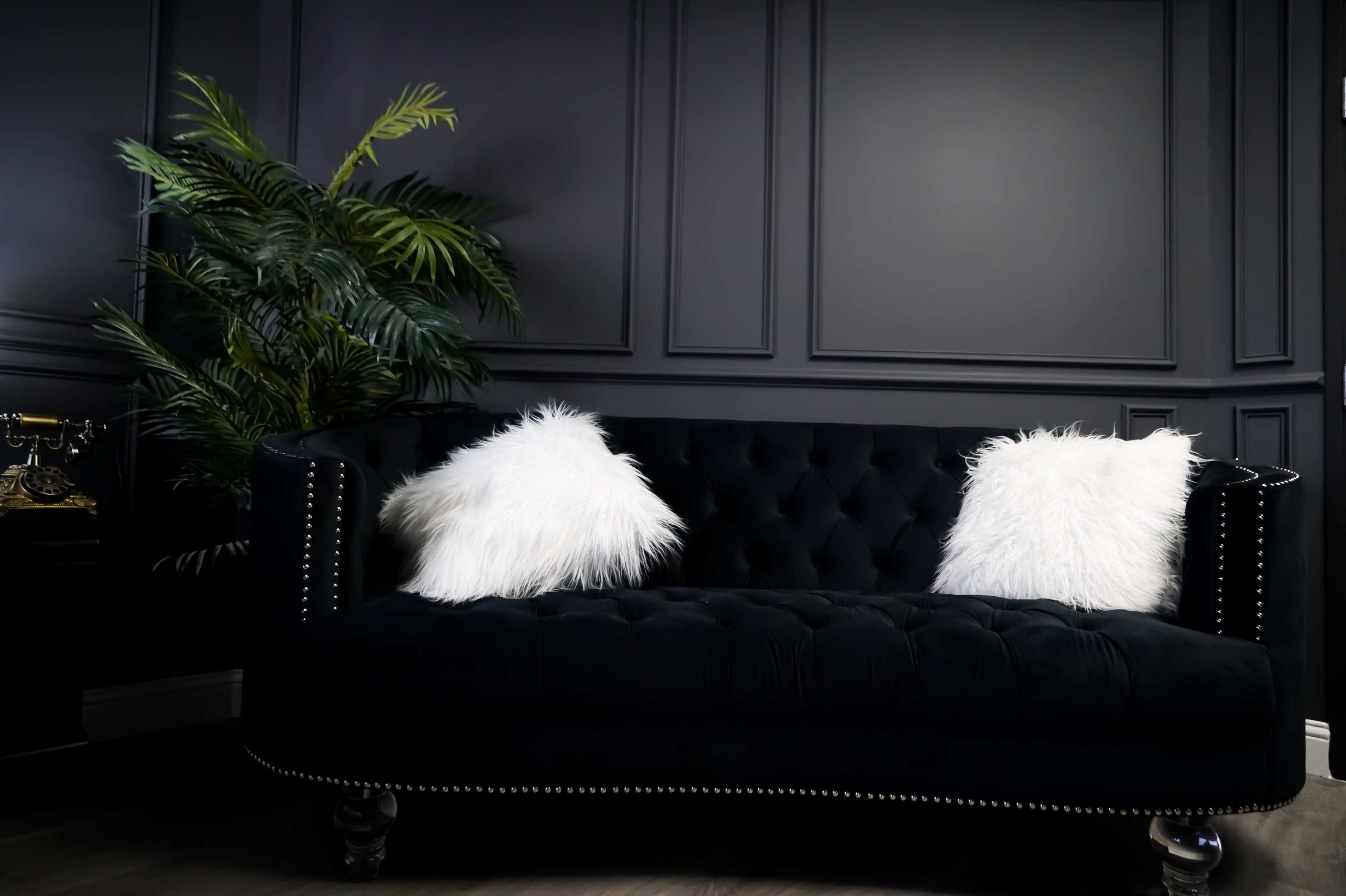 A black tufted sofa with white fur pillows is positioned against a dark paneled wall, with a large green plant nearby.