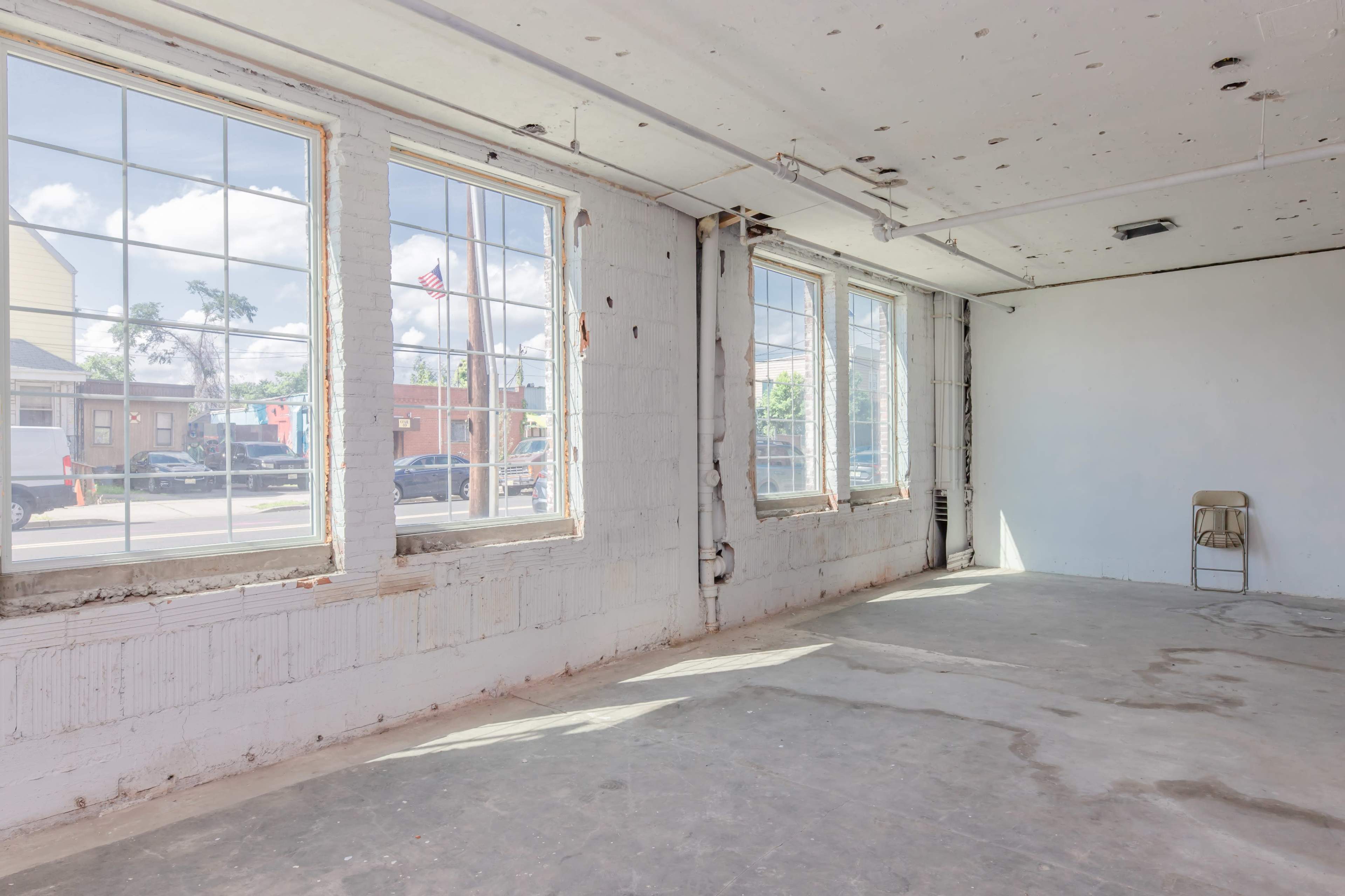 An empty room with large windows allows natural light to fill the space, featuring a single folding chair in one corner.