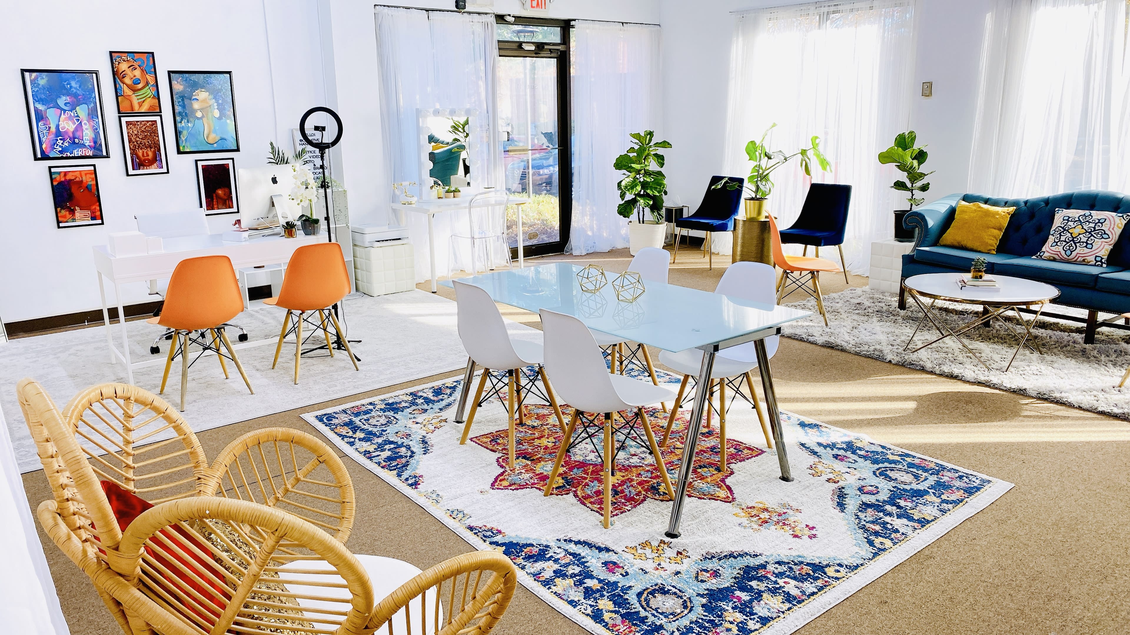 A brightly lit, modern interior featuring a dining table with white chairs, a blue sofa, various plants, and colorful artwork on the walls.