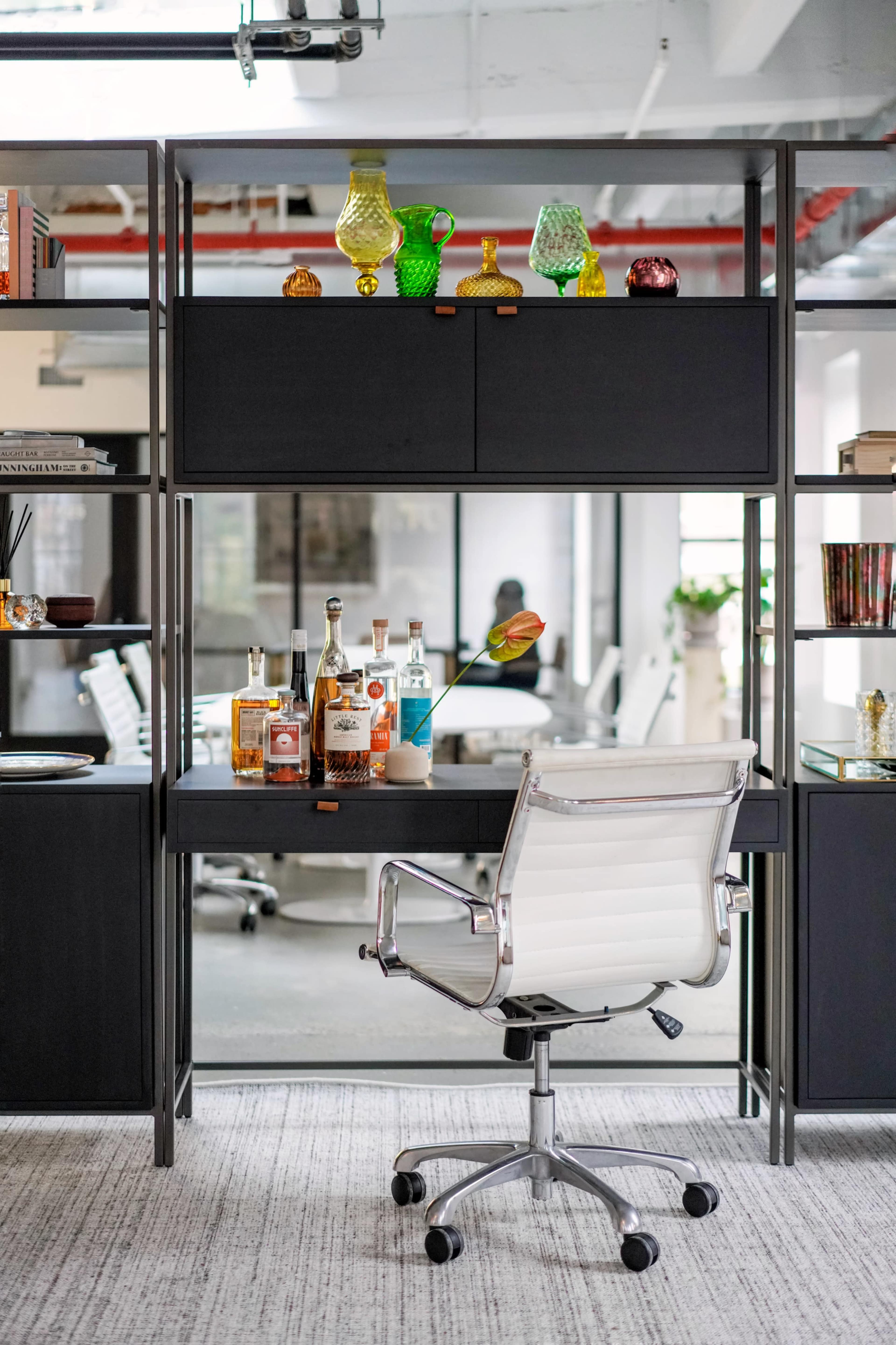 The image shows a modern desk setup in an office, featuring a chair, bottles on the desk, and decorative glass items on a shelf above.