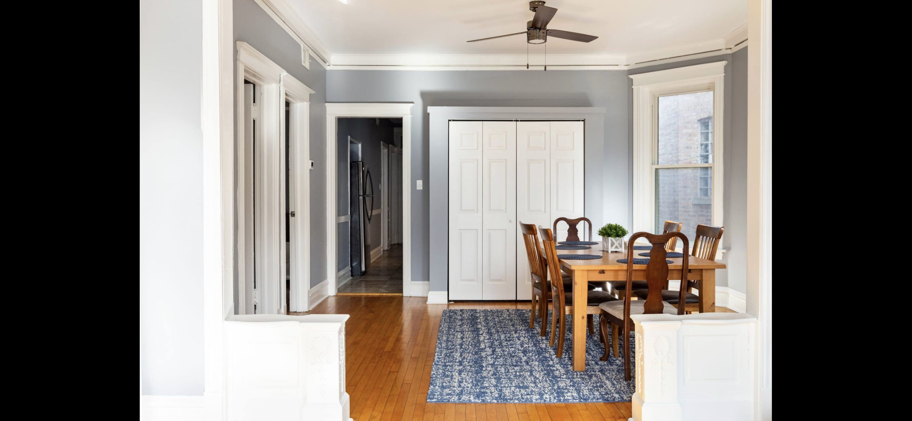 A dining area with a wooden table and chairs, situated on a blue rug, adjacent to light gray walls and a ceiling fan.