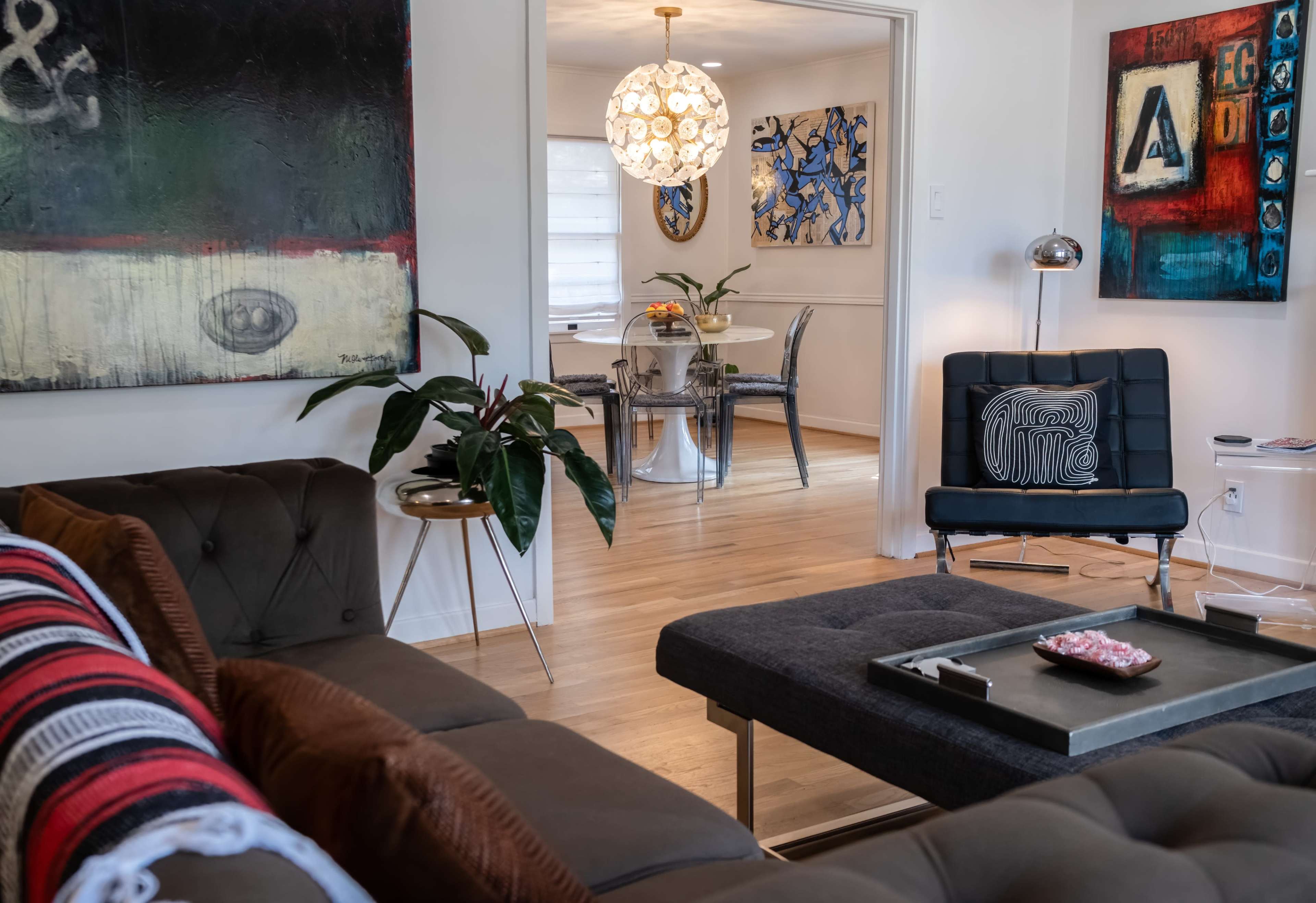 The image shows a modern living room with a dark sofa, a coffee table, and a dining area visible through an open doorway, decorated with artwork and lighting.