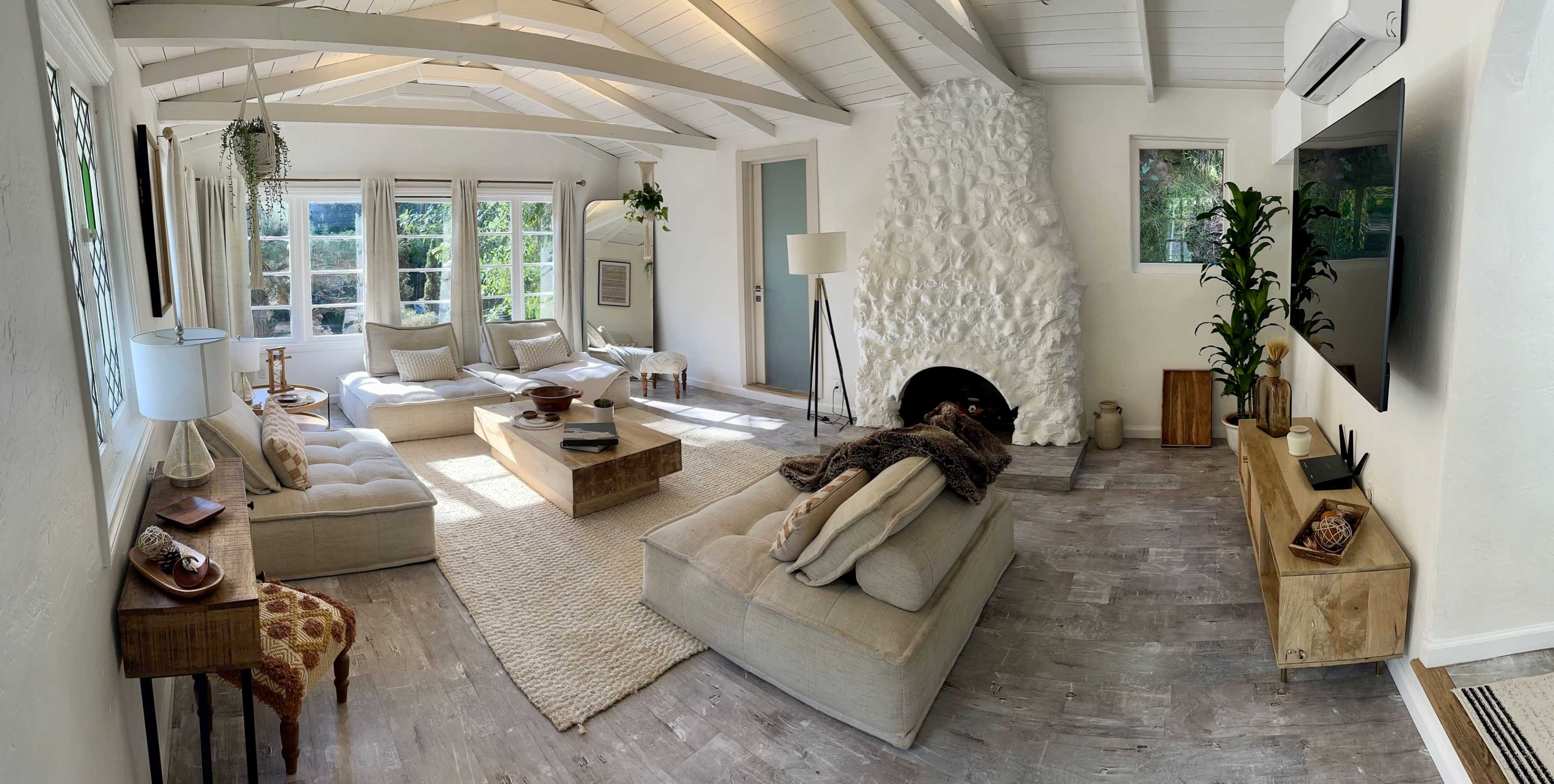 A bright living room with a white stone fireplace, large windows, and beige furniture arranged around a wooden coffee table.
