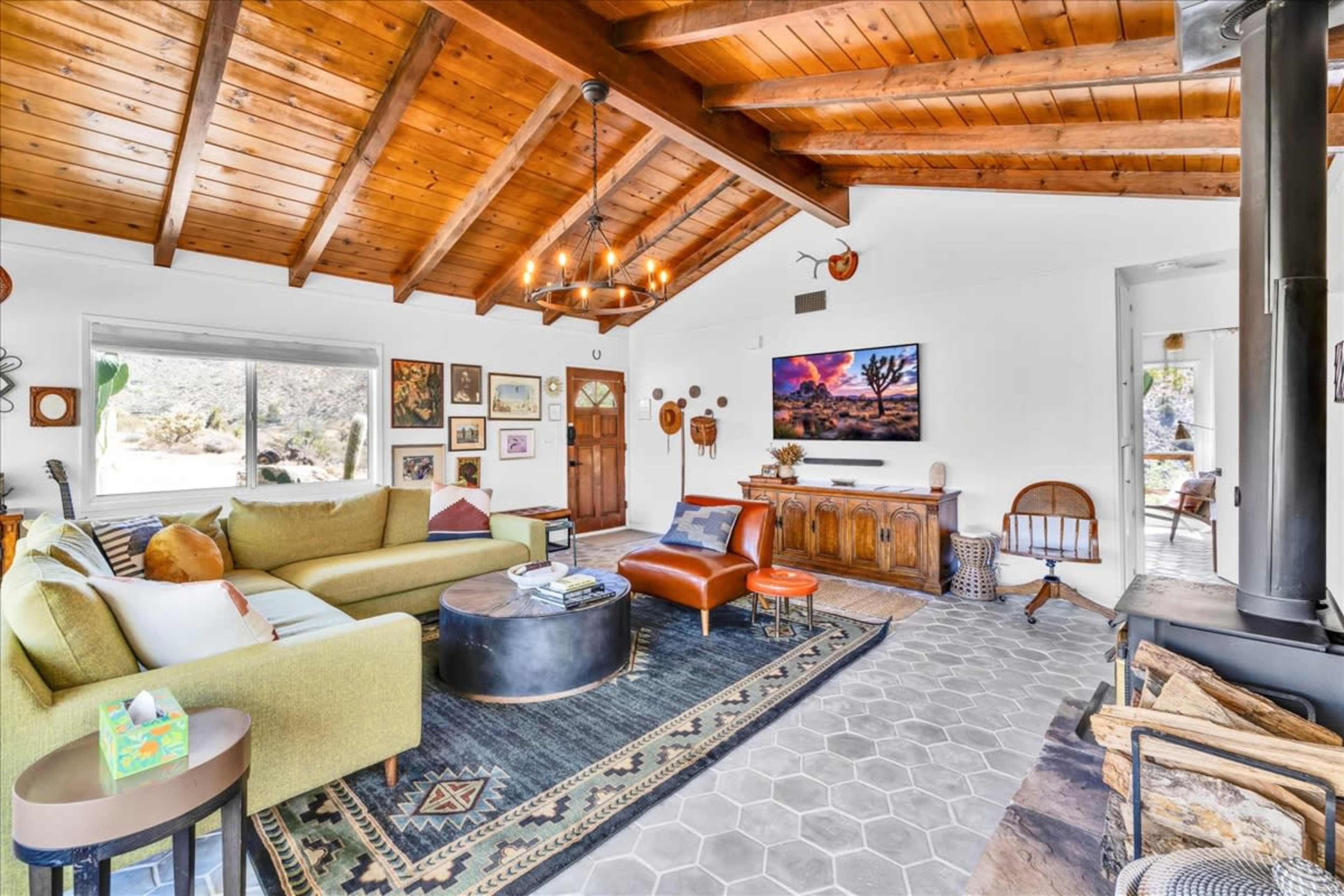 A cozy living room features a wooden beam ceiling, a large green sectional sofa, and a fireplace, surrounded by various artworks and decor.