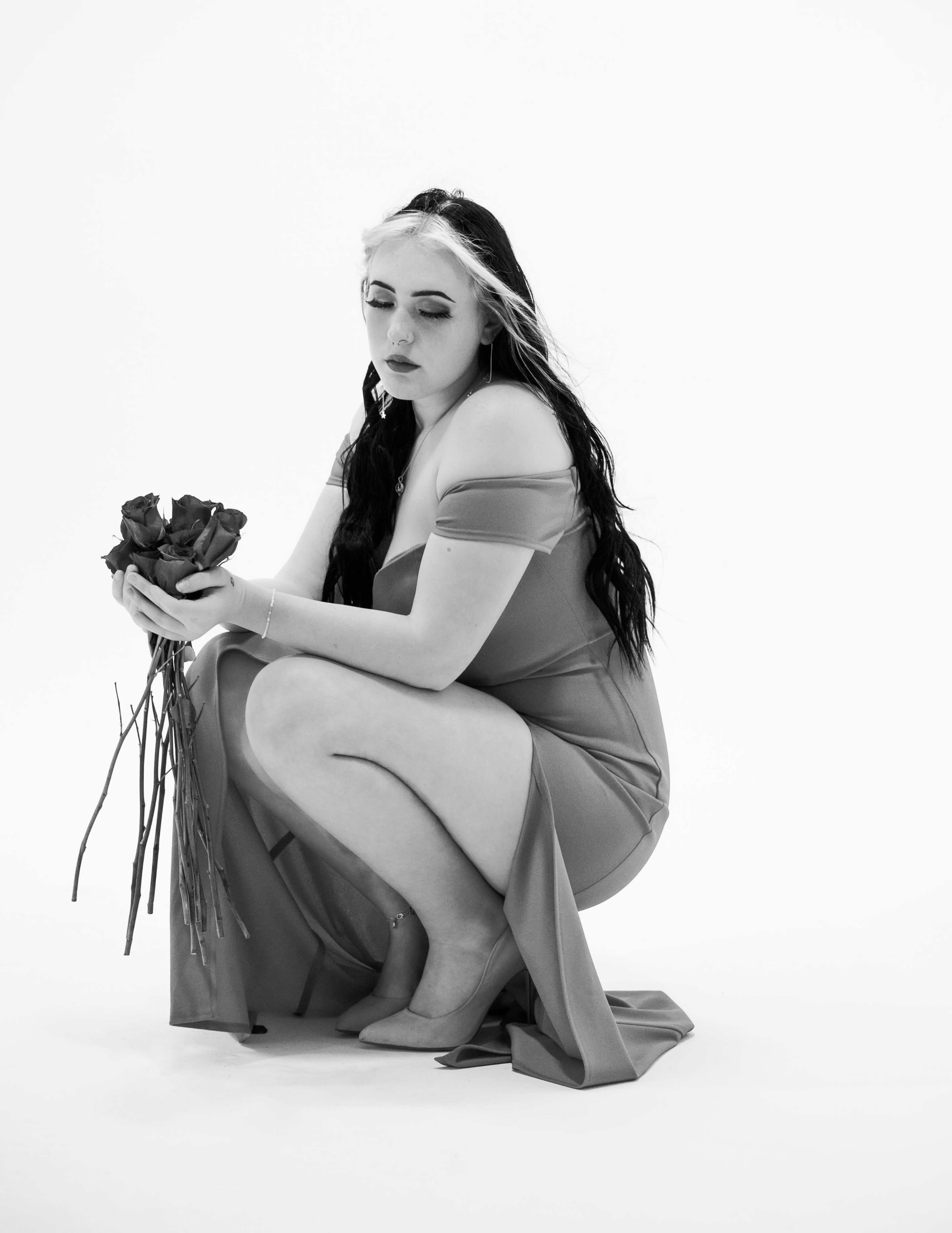 A woman in a long, flowing dress squats on the floor while holding a bouquet of roses.