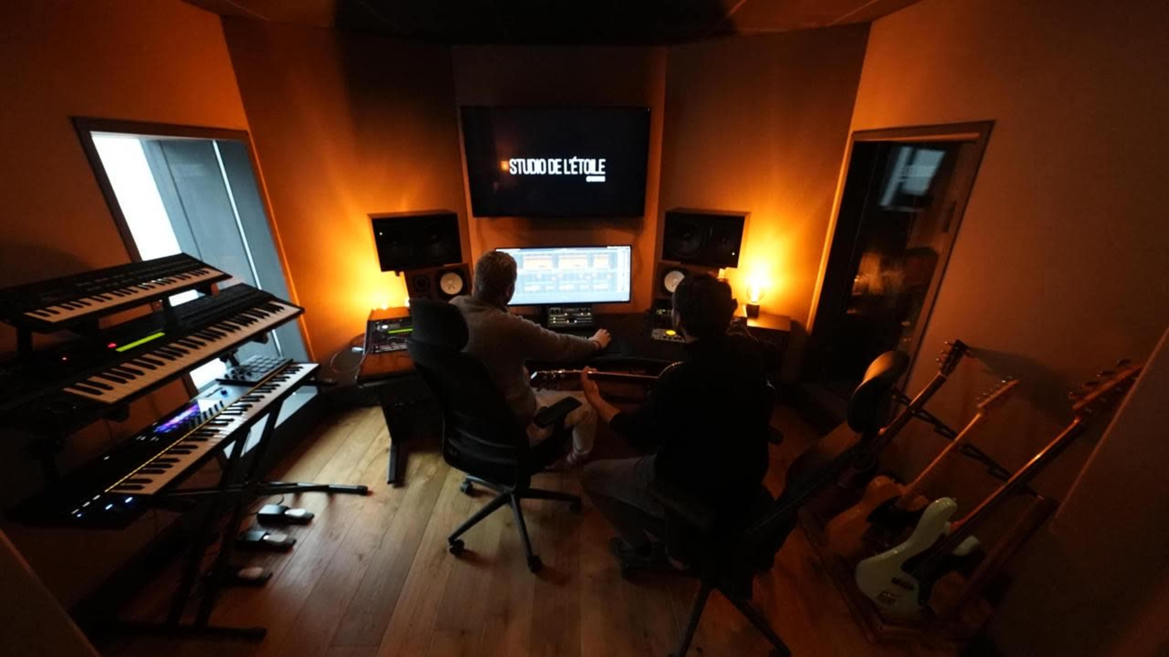 Two individuals are working together in a music studio surrounded by electronic instruments, mixing equipment, and warm lighting.