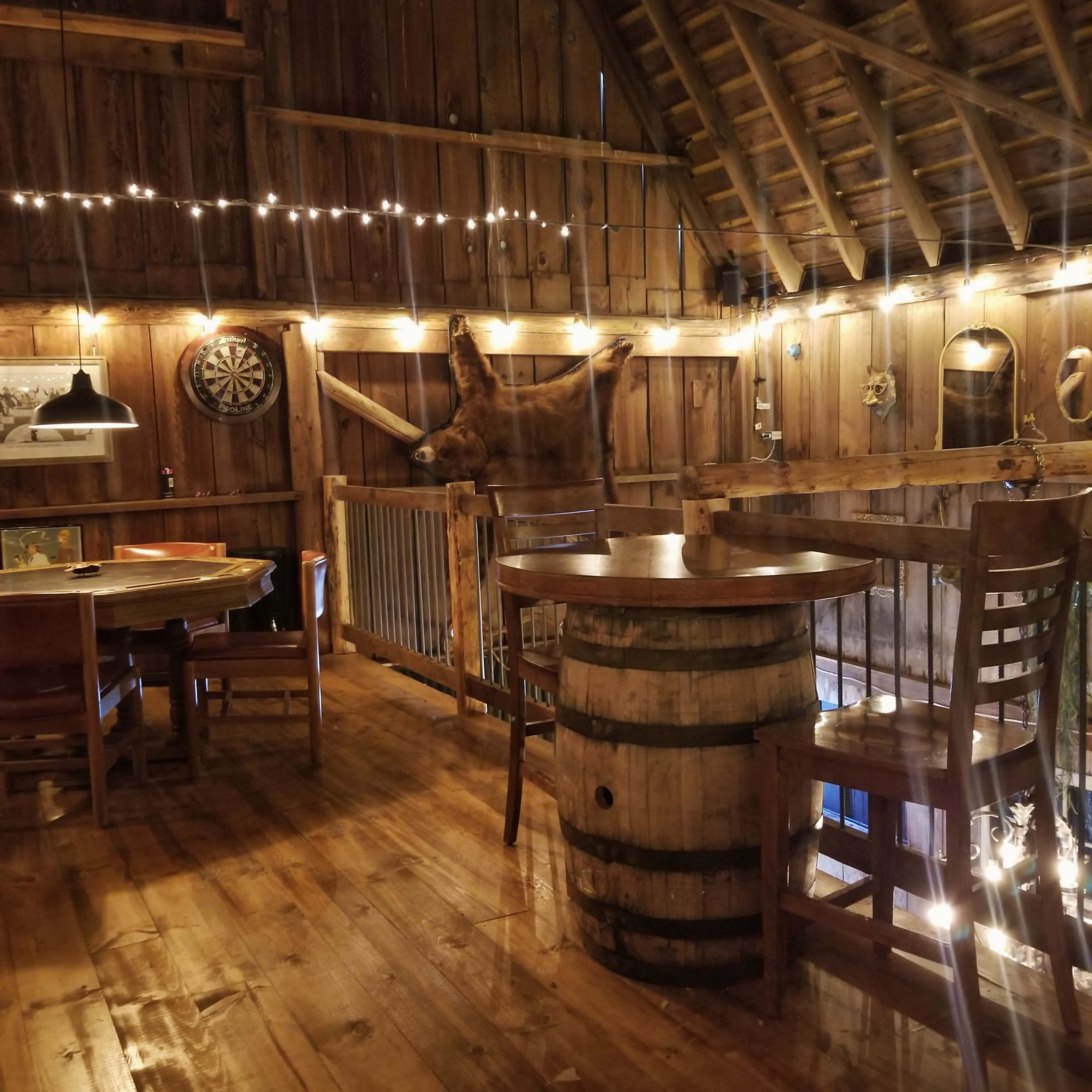 The interior of a rustic barn features wooden walls, a mounted animal head, a round barrel table, and string lights overhead.