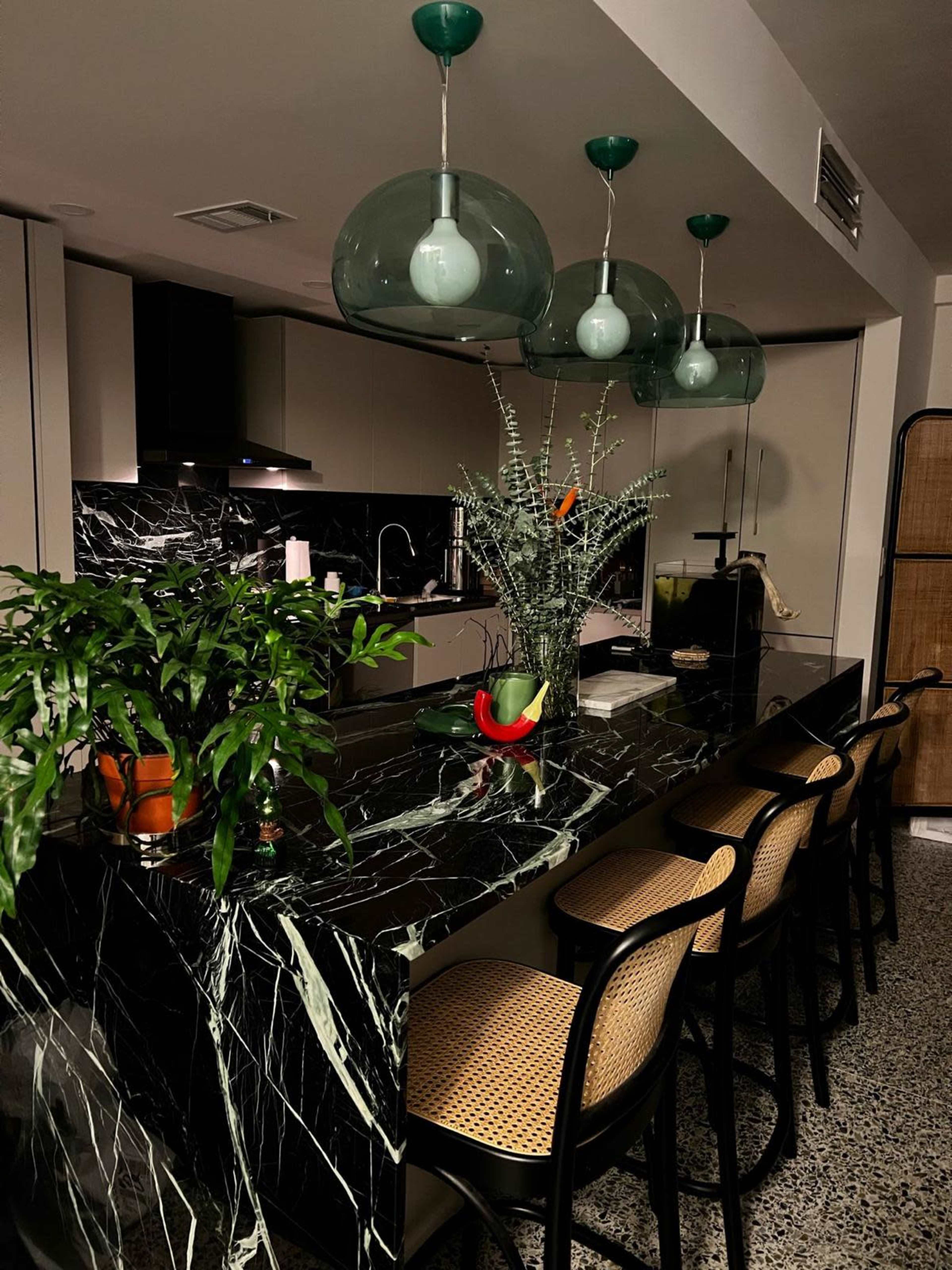 A modern kitchen featuring a black marble countertop, three green pendant lights, and bar stools along the counter, with plants and a fruit bowl as decor.