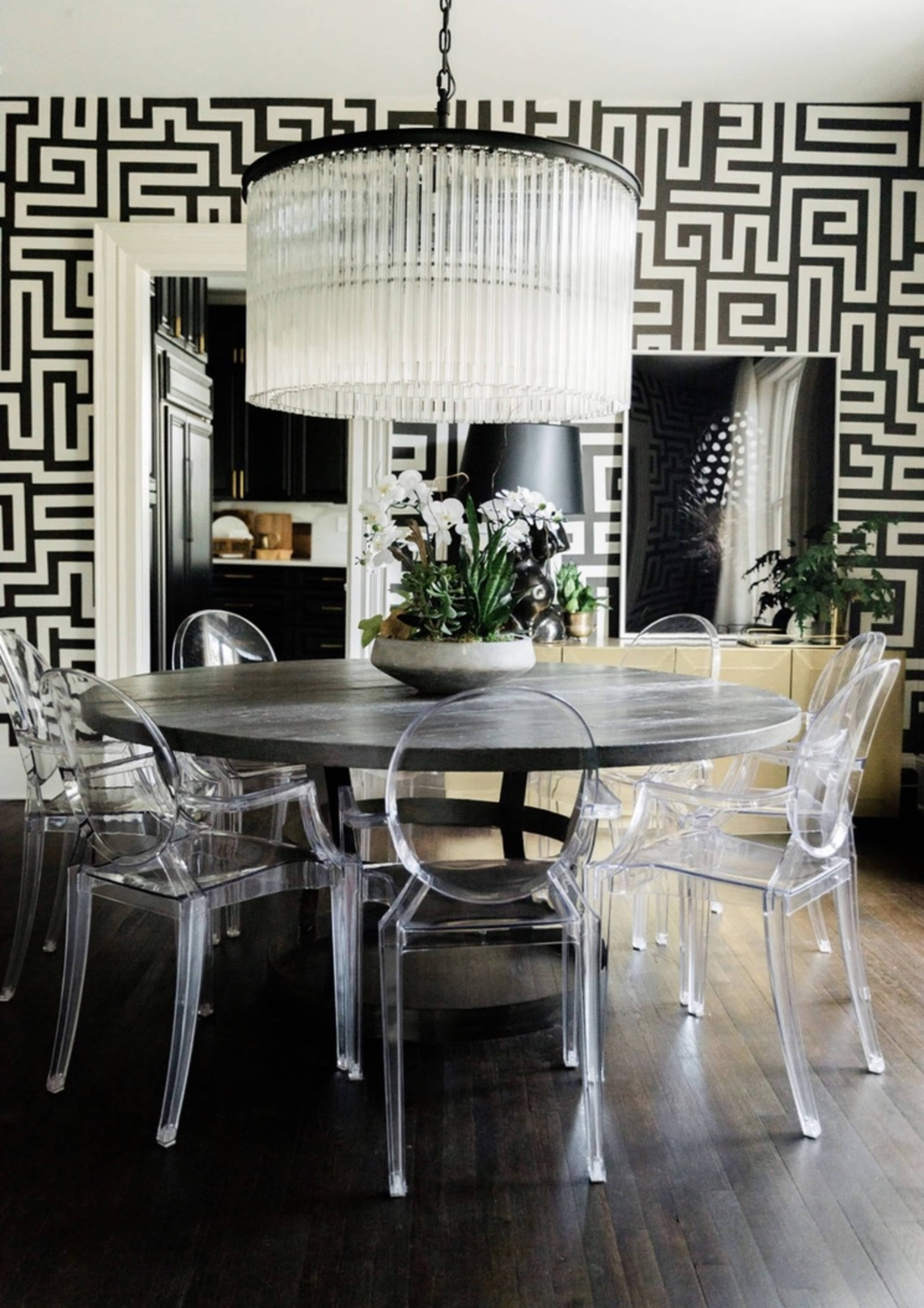 A round dining table surrounded by clear acrylic chairs sits under a large, ornate chandelier in a room with a patterned black and white wallpaper.