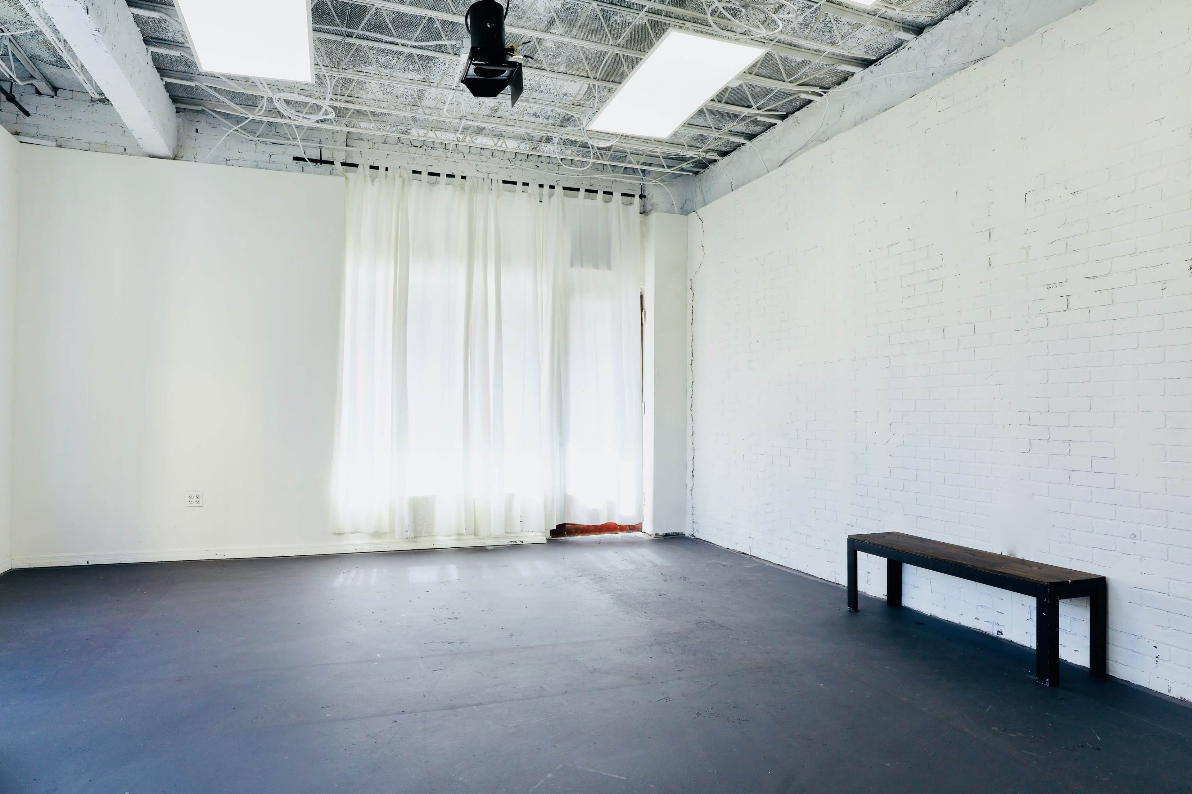 A minimalist, empty room with white walls, a wooden bench, and large curtains near a window.