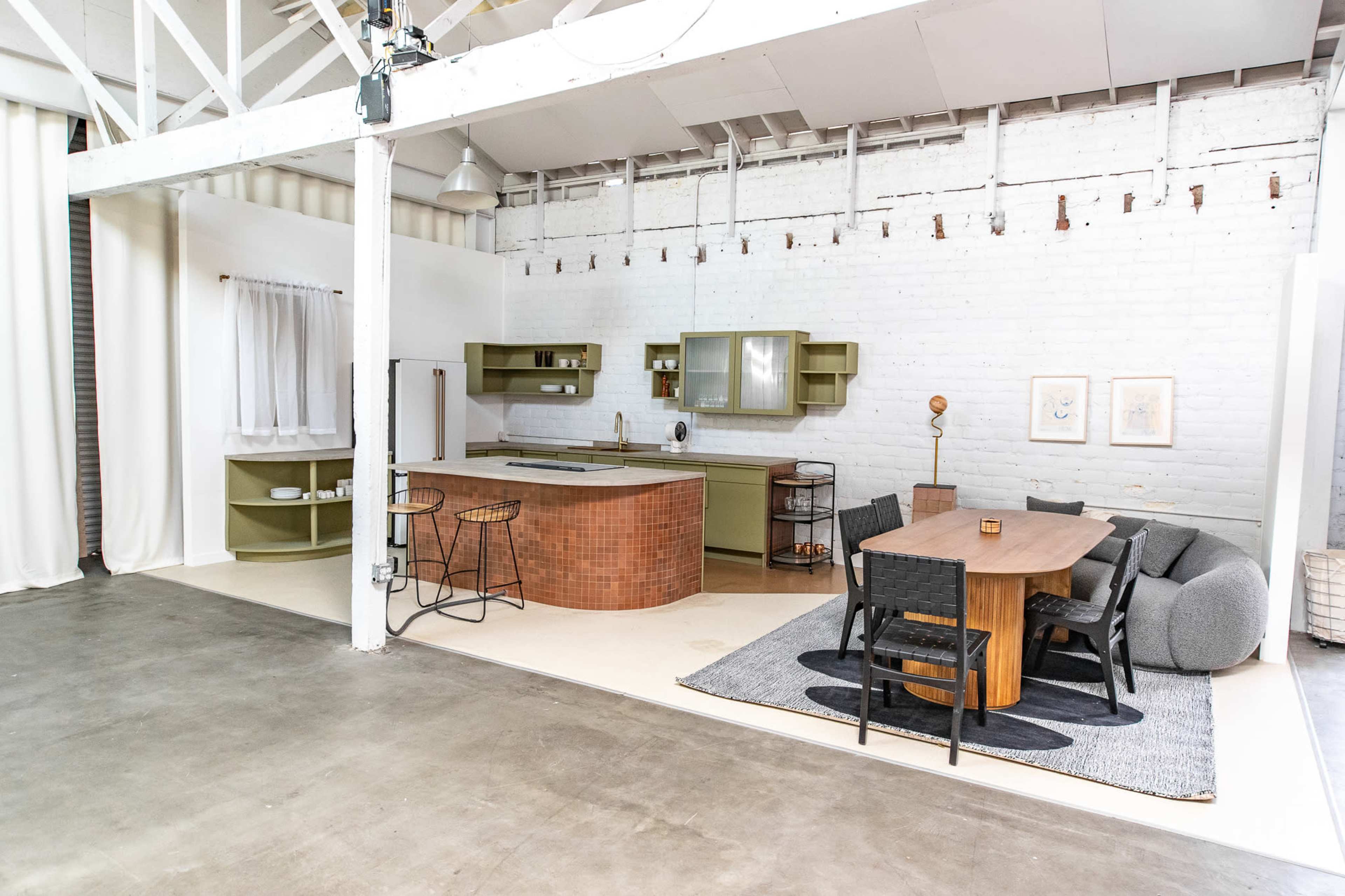 A spacious, modern kitchen and dining area featuring a large wooden table, black chairs, and a kitchen island with green cabinetry against a white brick wall.