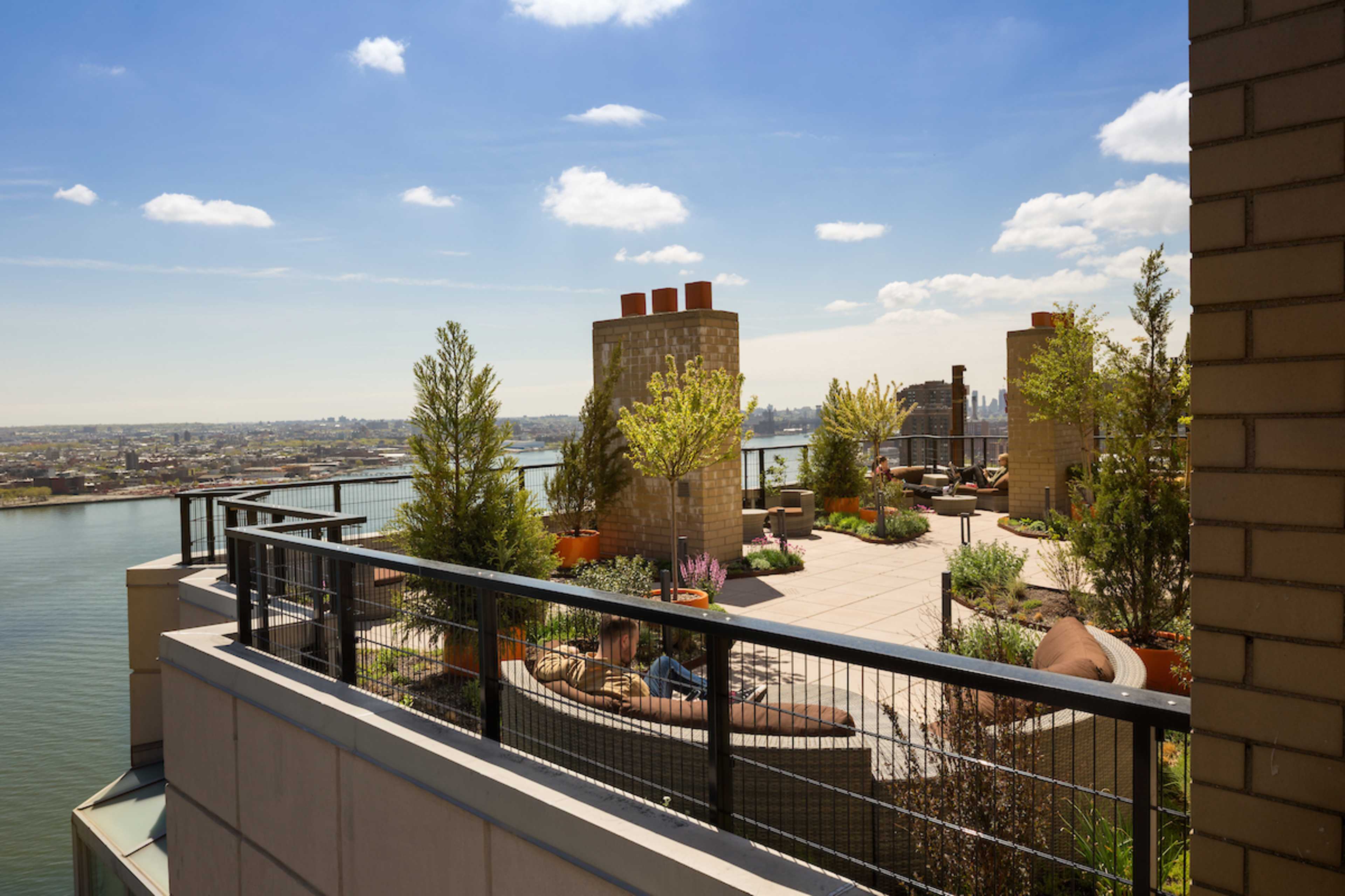 A rooftop terrace with seating and potted plants overlooking a river and cityscape under a blue sky.