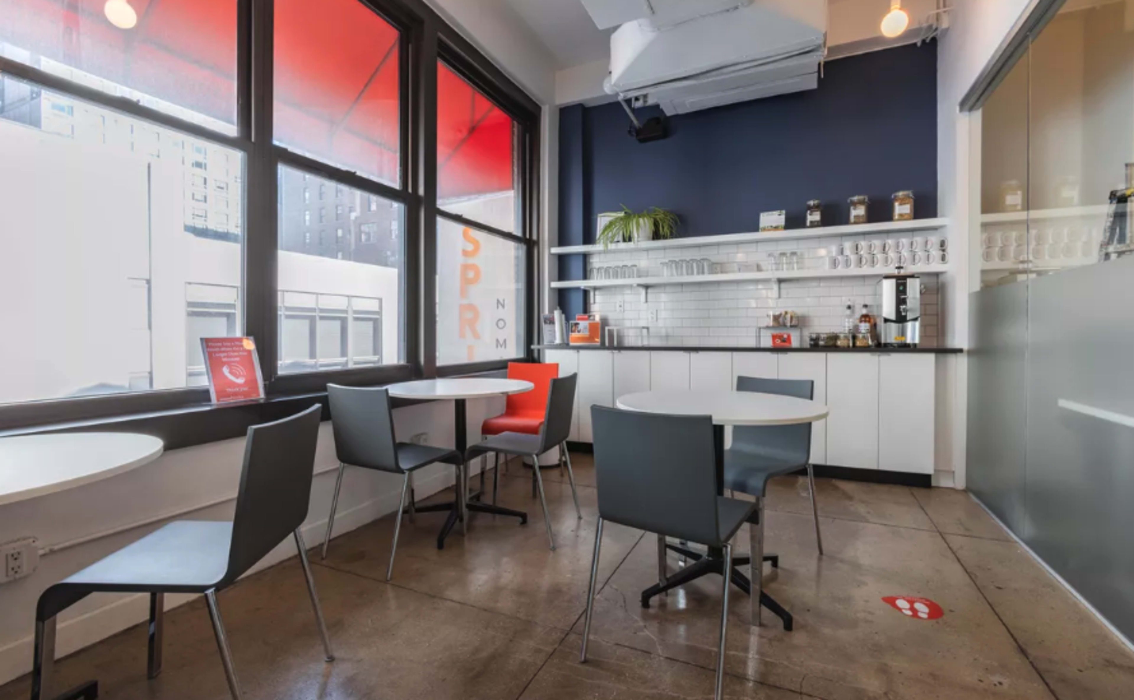 A modern, minimalist café space with gray chairs, round tables, and large windows featuring red shades.