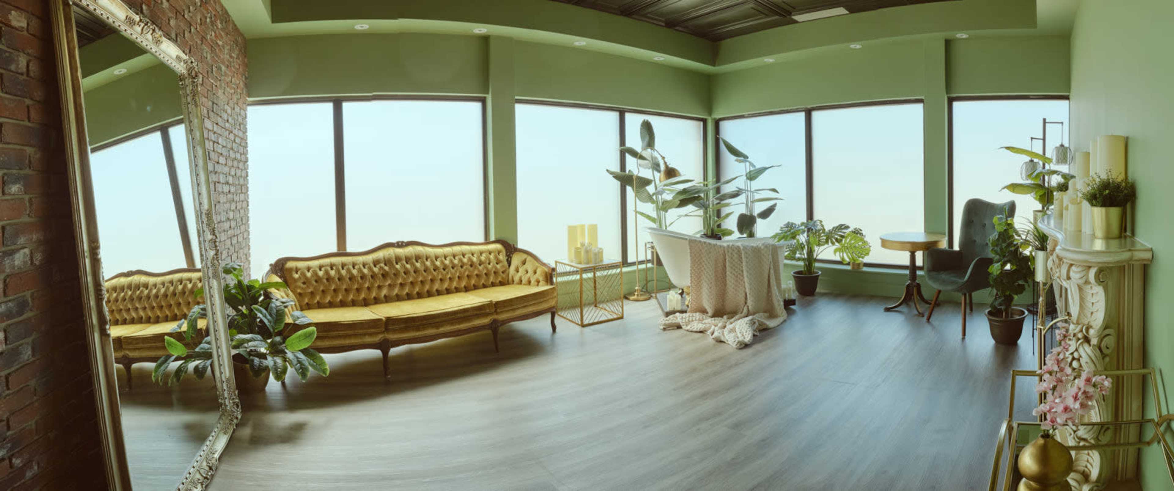 The image shows a spacious, modern living room with large windows, a golden couch, various plants, and elegant decor elements.