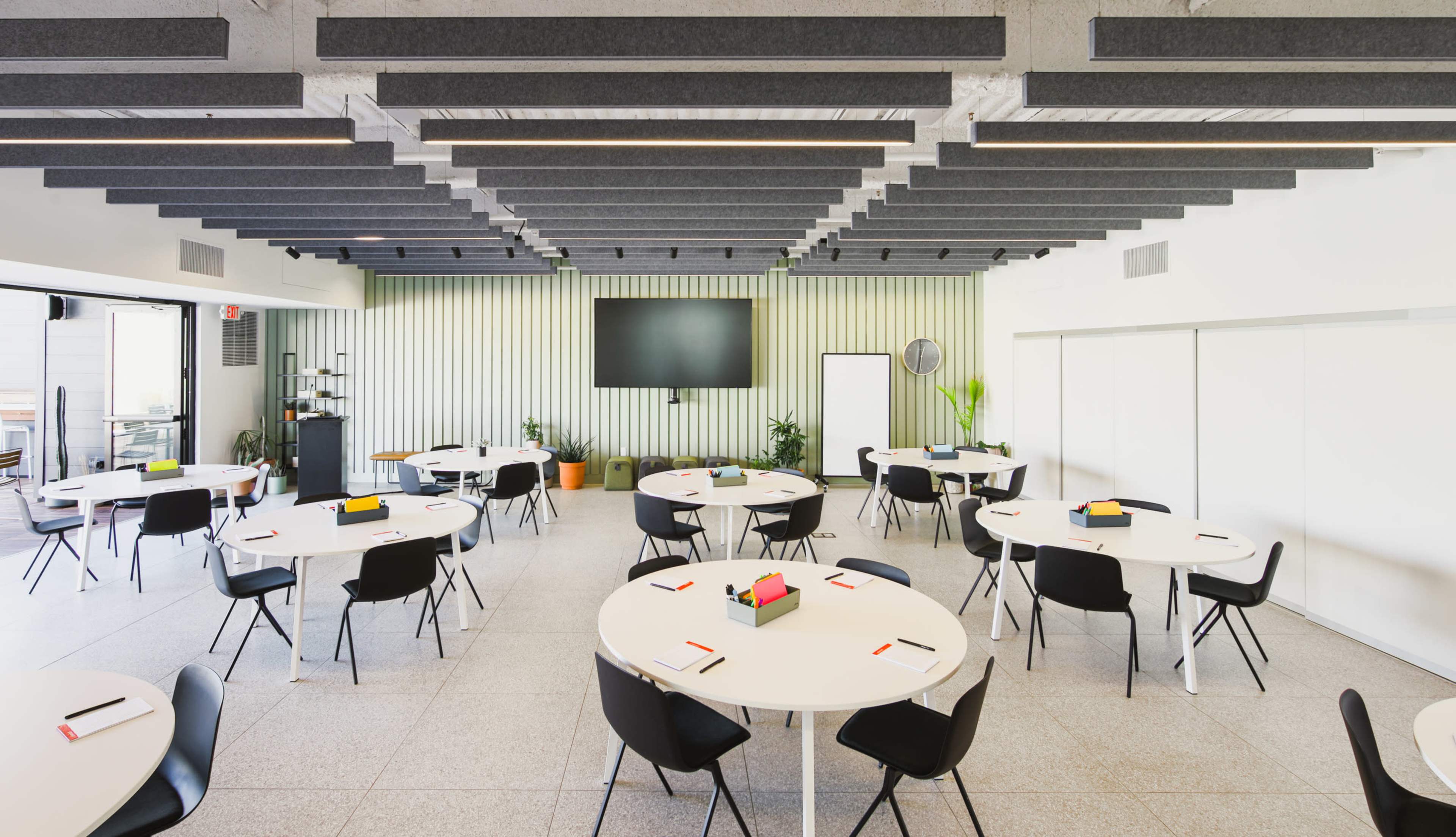 A modern conference room features multiple round tables set with stationery, surrounded by sleek black chairs and equipped with a large screen at one end.