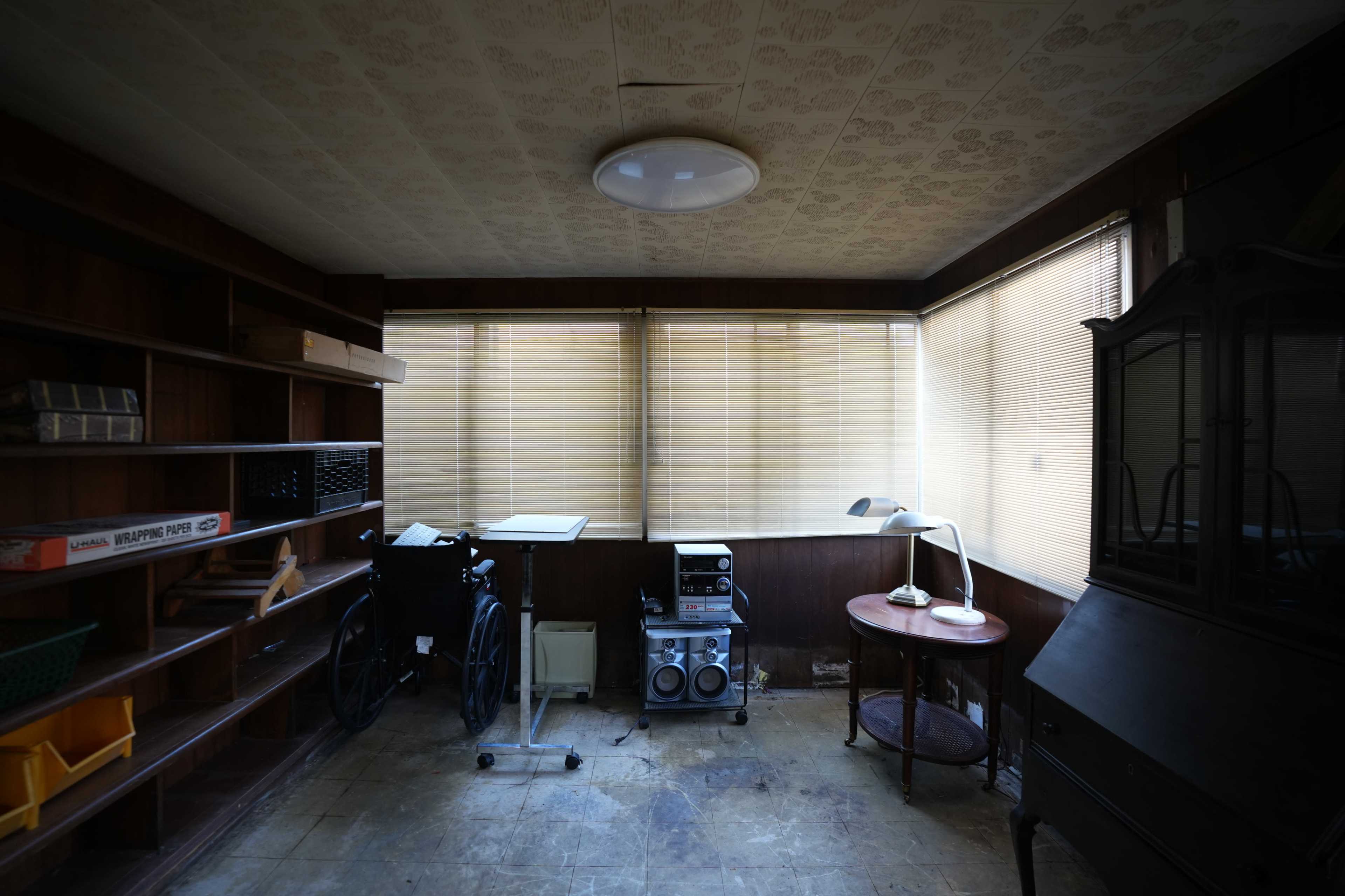The image shows an empty room with wooden shelves, a wheelchair, a small table with a lamp, and a stereo system, illuminated by natural light through partially covered windows.