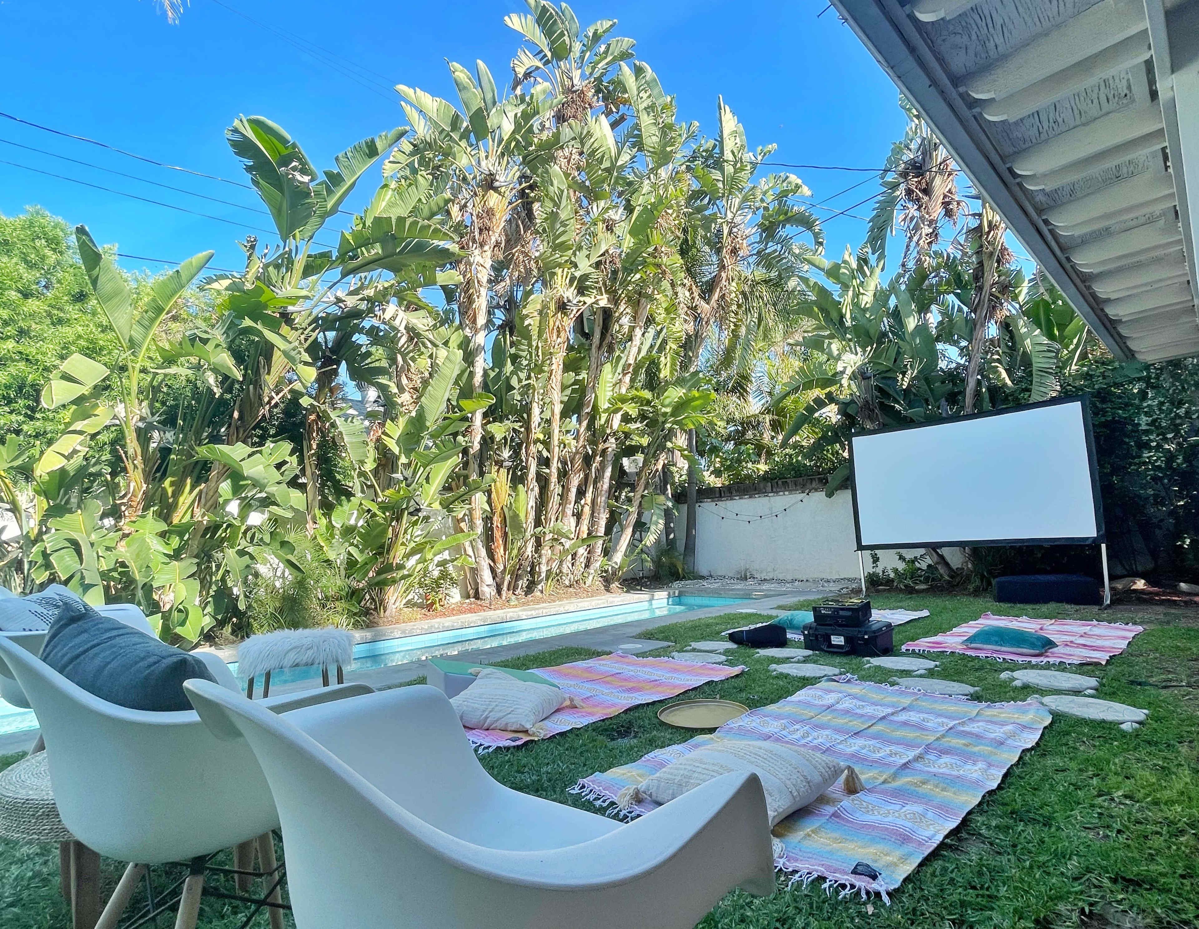 A backyard setup features white chairs and colorful blankets around a projector screen beside a swimming pool, surrounded by lush green palm trees.
