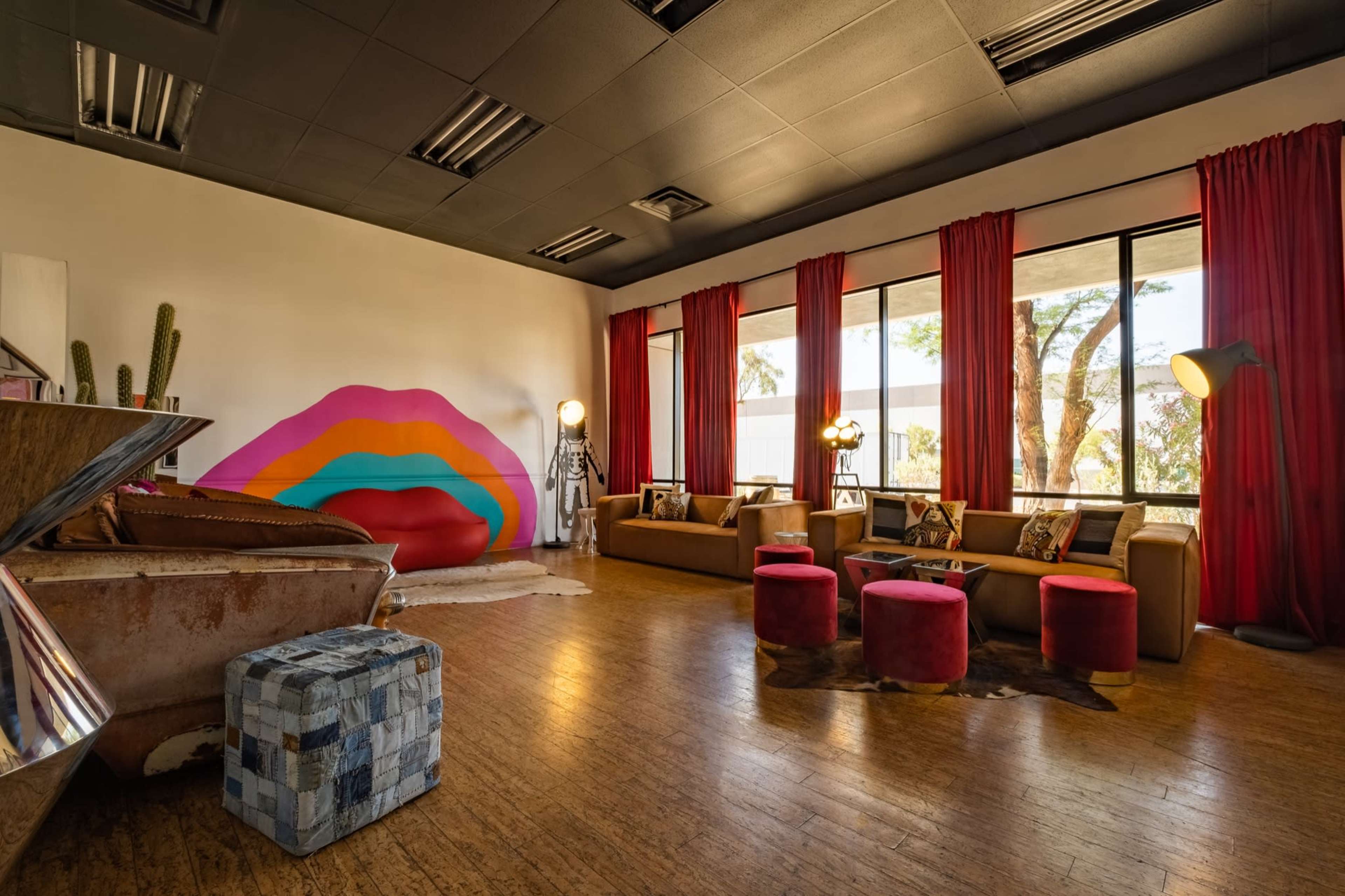 The image shows a brightly lit lounge area featuring a large, colorful wall mural, modern furniture, and floor-to-ceiling windows with red curtains.