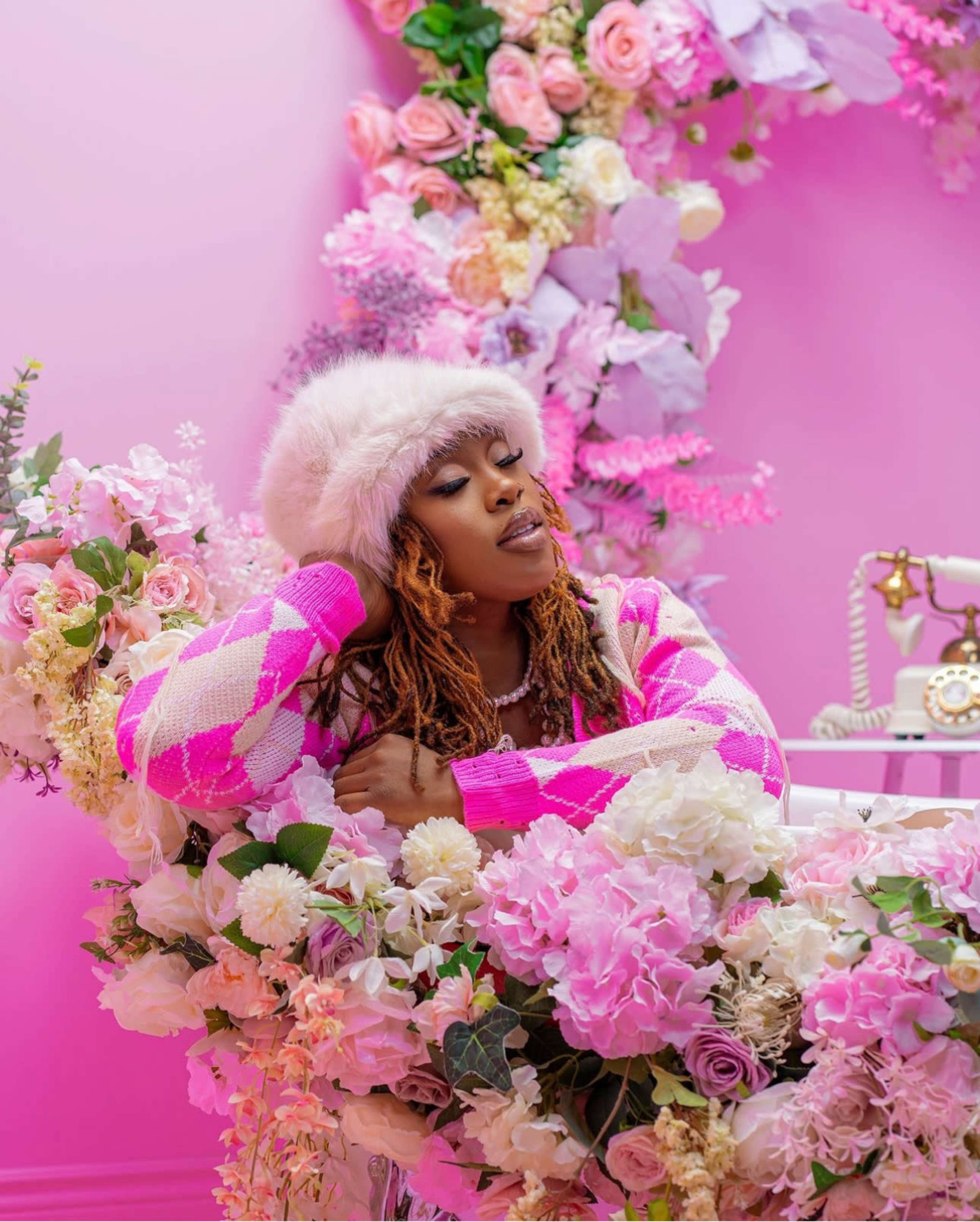 A person poses amid a decorative arrangement of pink flowers and a vintage telephone against a pink backdrop.