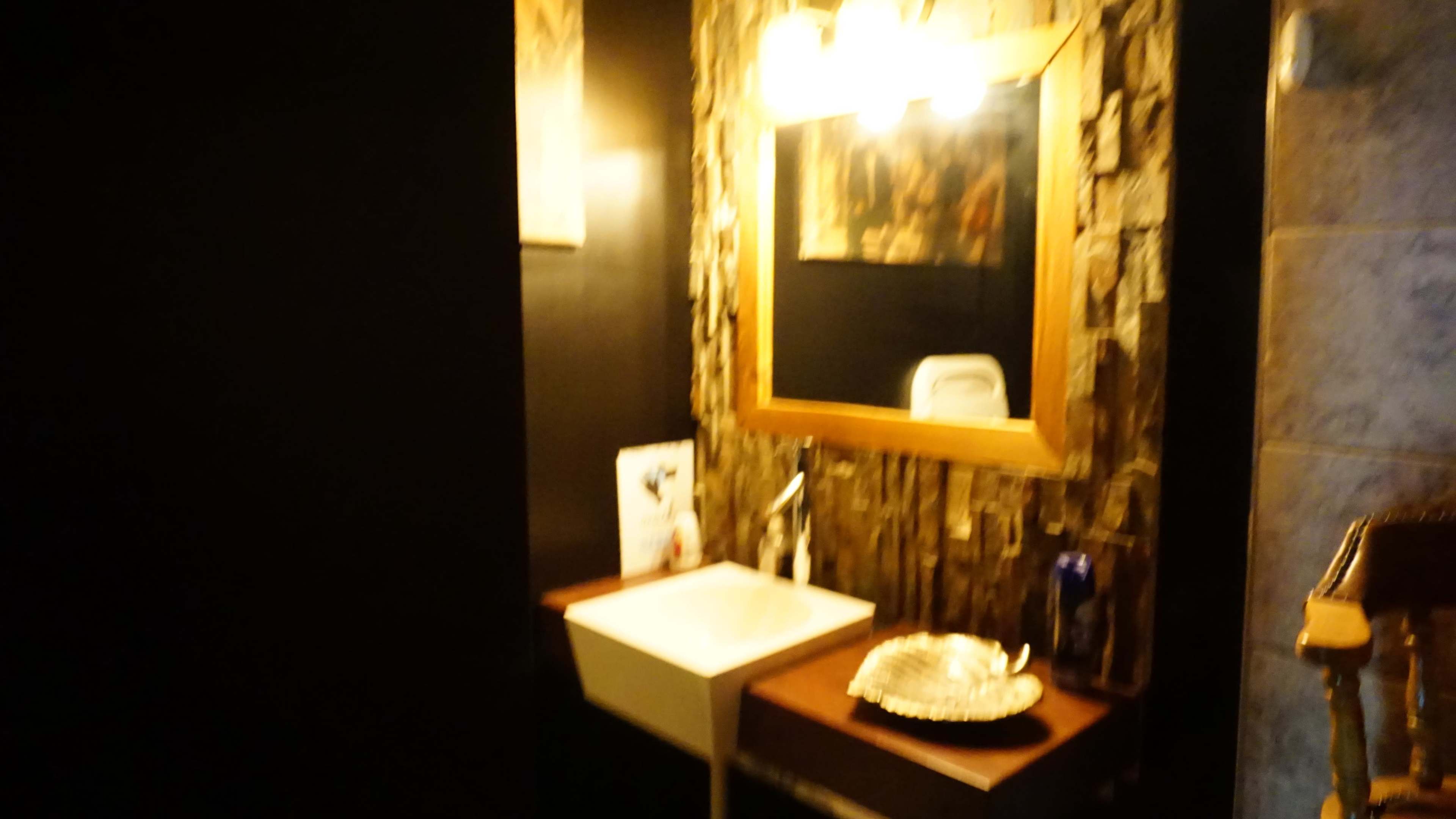 The image shows a small bathroom with a stone-patterned wall, a rectangular sink, a wooden mirror frame, and soft lighting.