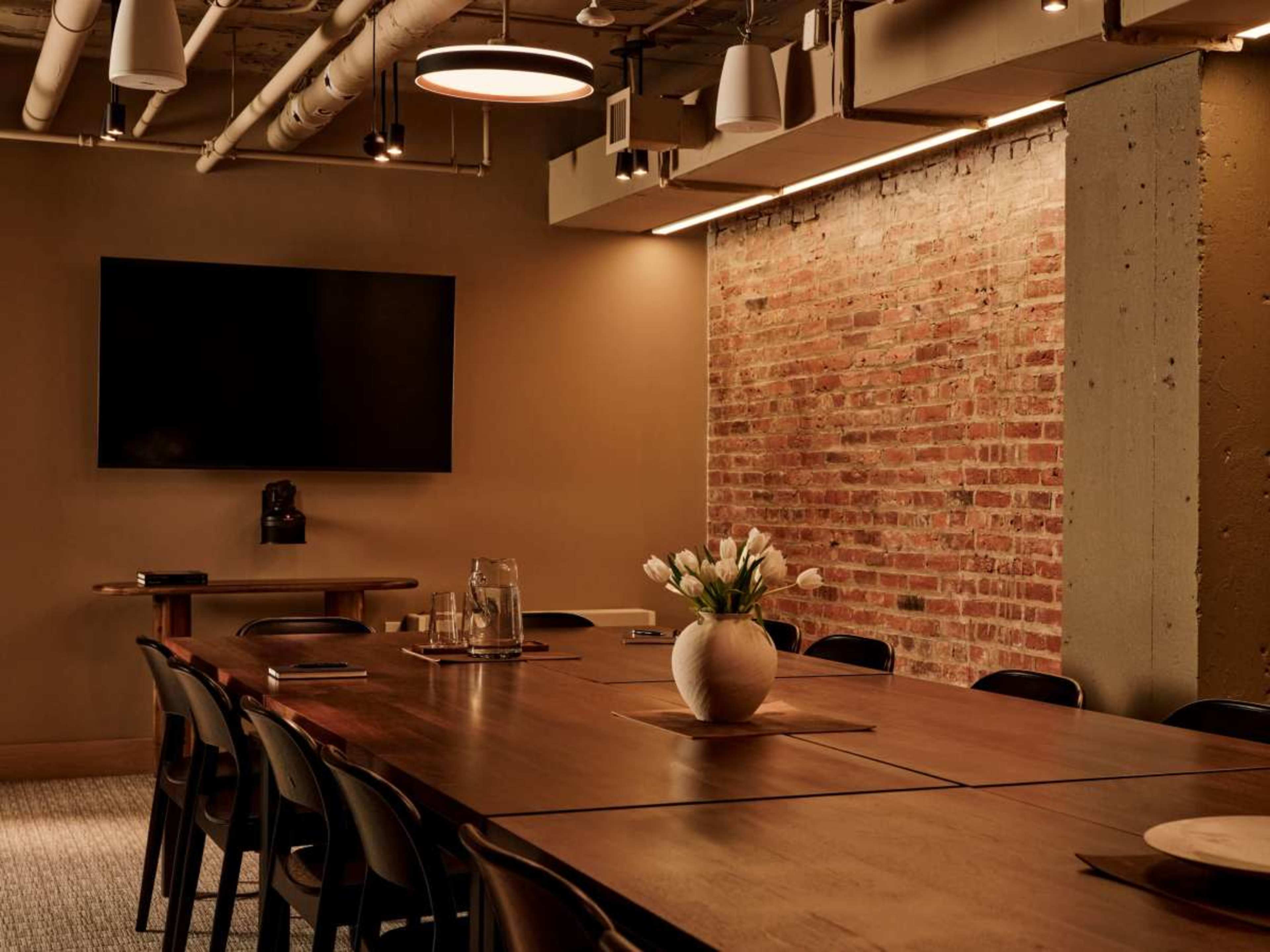 A modern conference room features a large wooden table with a vase of white tulips at its center, exposed brick walls, and a wall-mounted television.