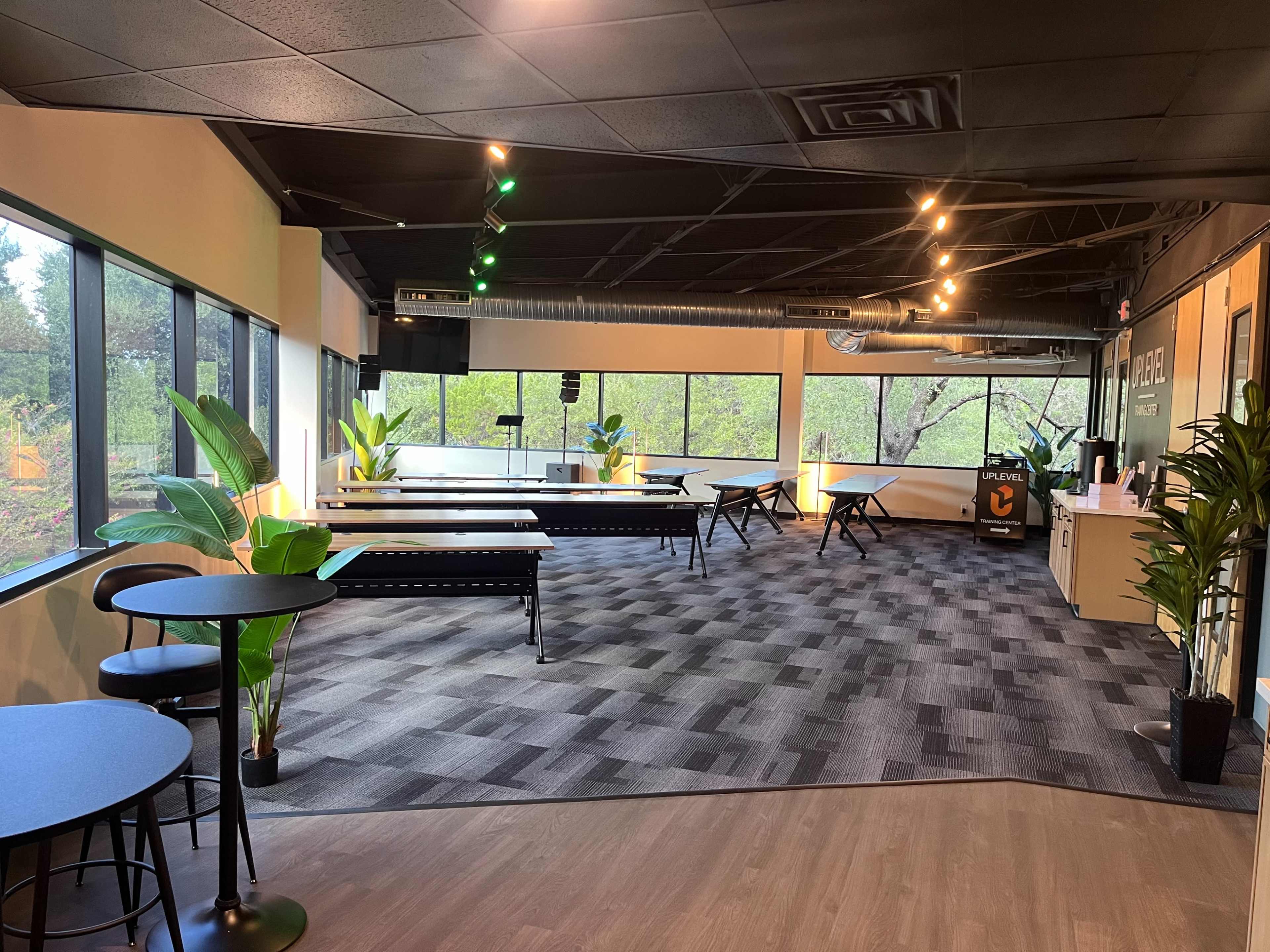 The image shows a spacious, well-lit office area with large windows, showcasing a gray carpet, black tables, and green plants scattered throughout.