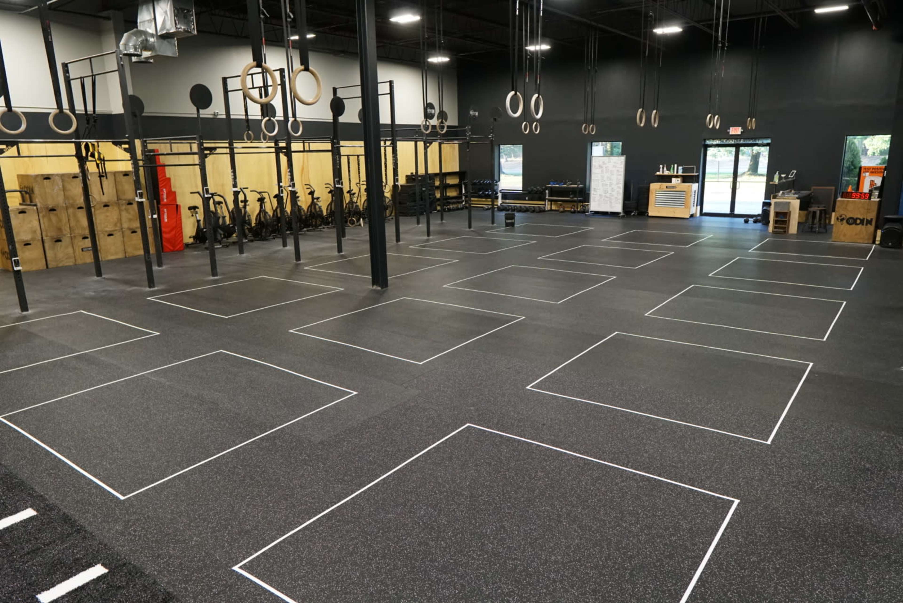 The image shows a spacious gym interior with marked workout areas, exercise equipment, and gymnastic rings hanging from the ceiling.