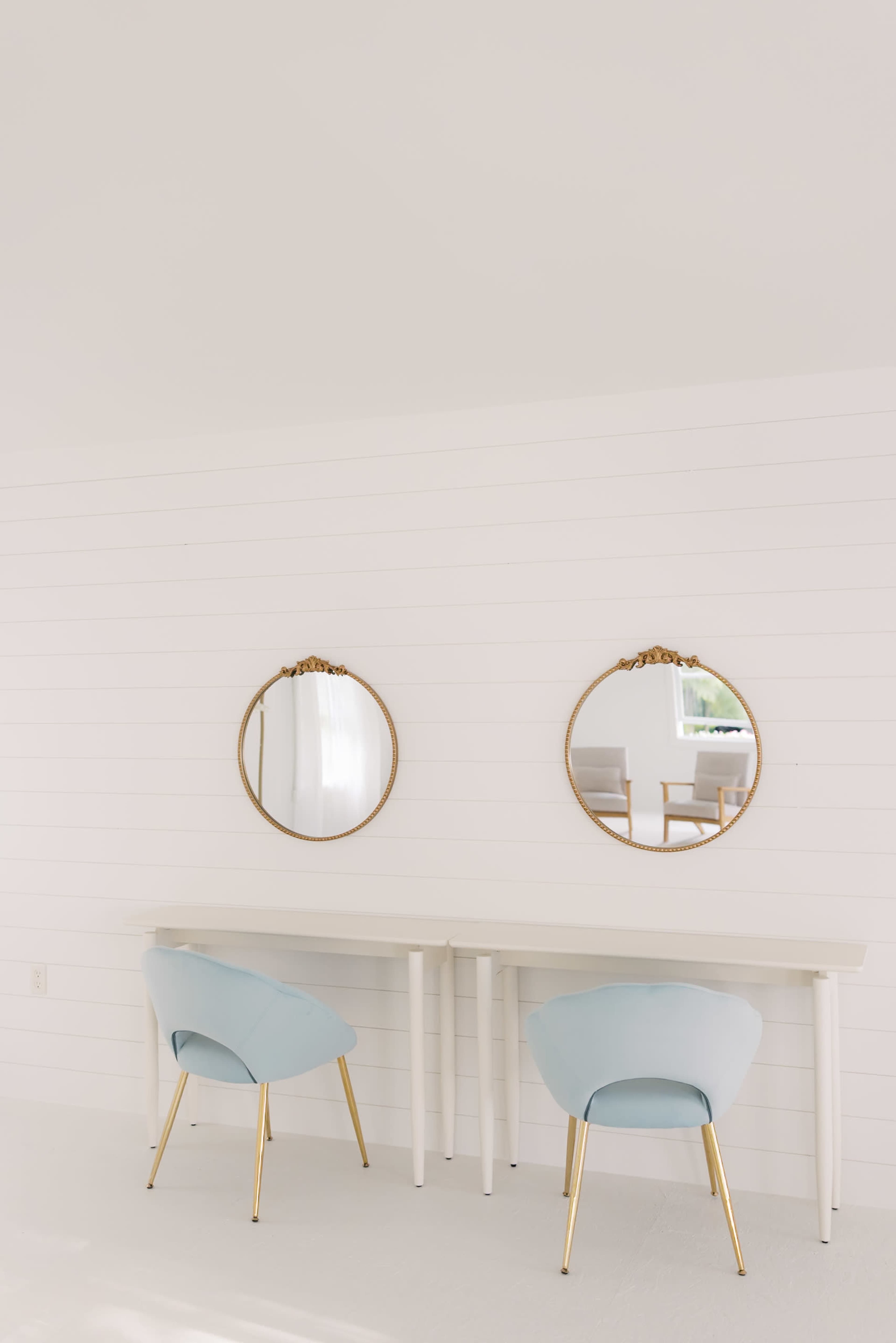 The image shows a minimalist white room featuring a long table with two round mirrors above it and two light blue chairs with gold legs.