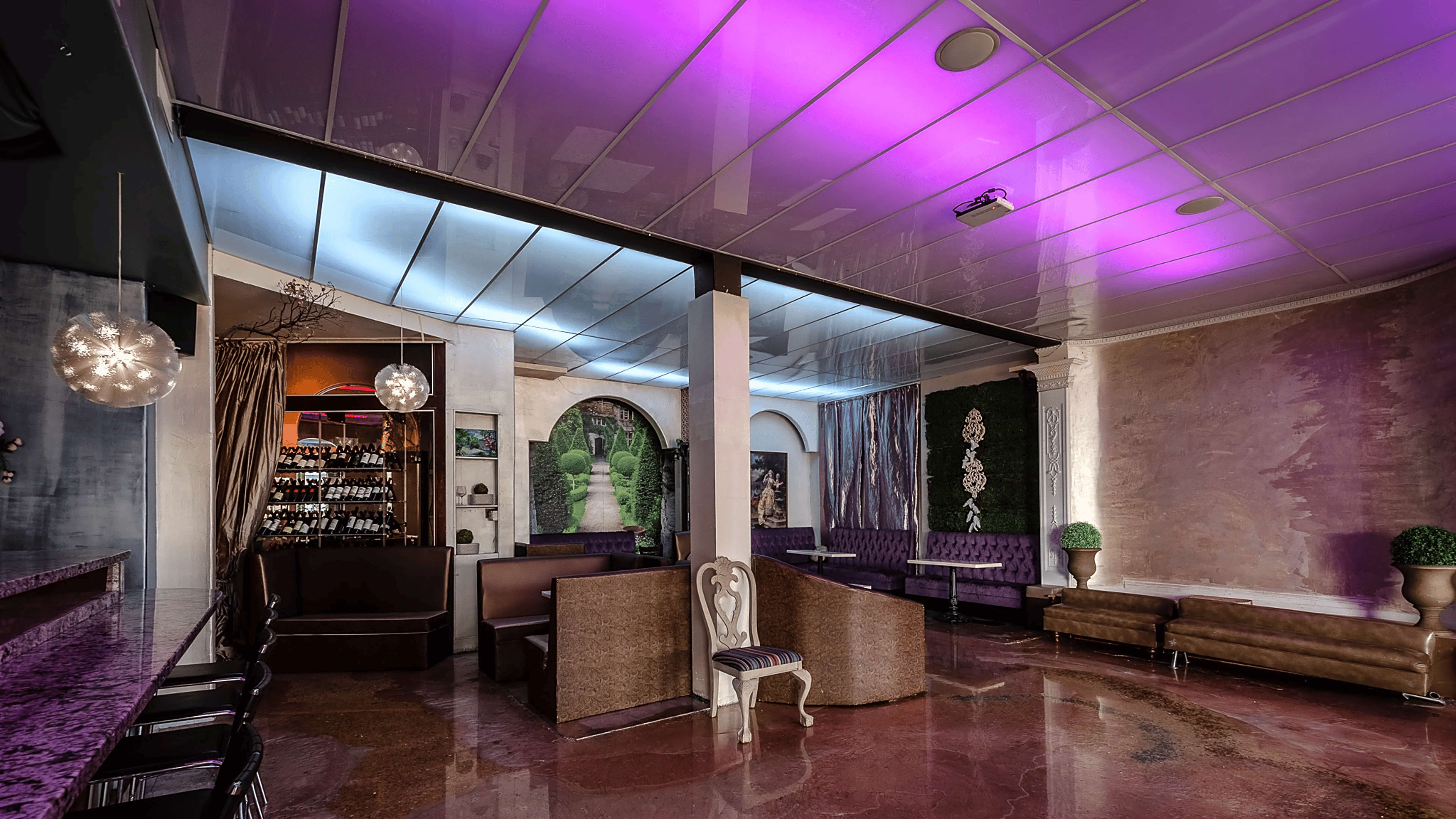 The image shows a modern lounge interior featuring plush seating, a bar with a wine display, and ambient lighting in shades of purple and blue.